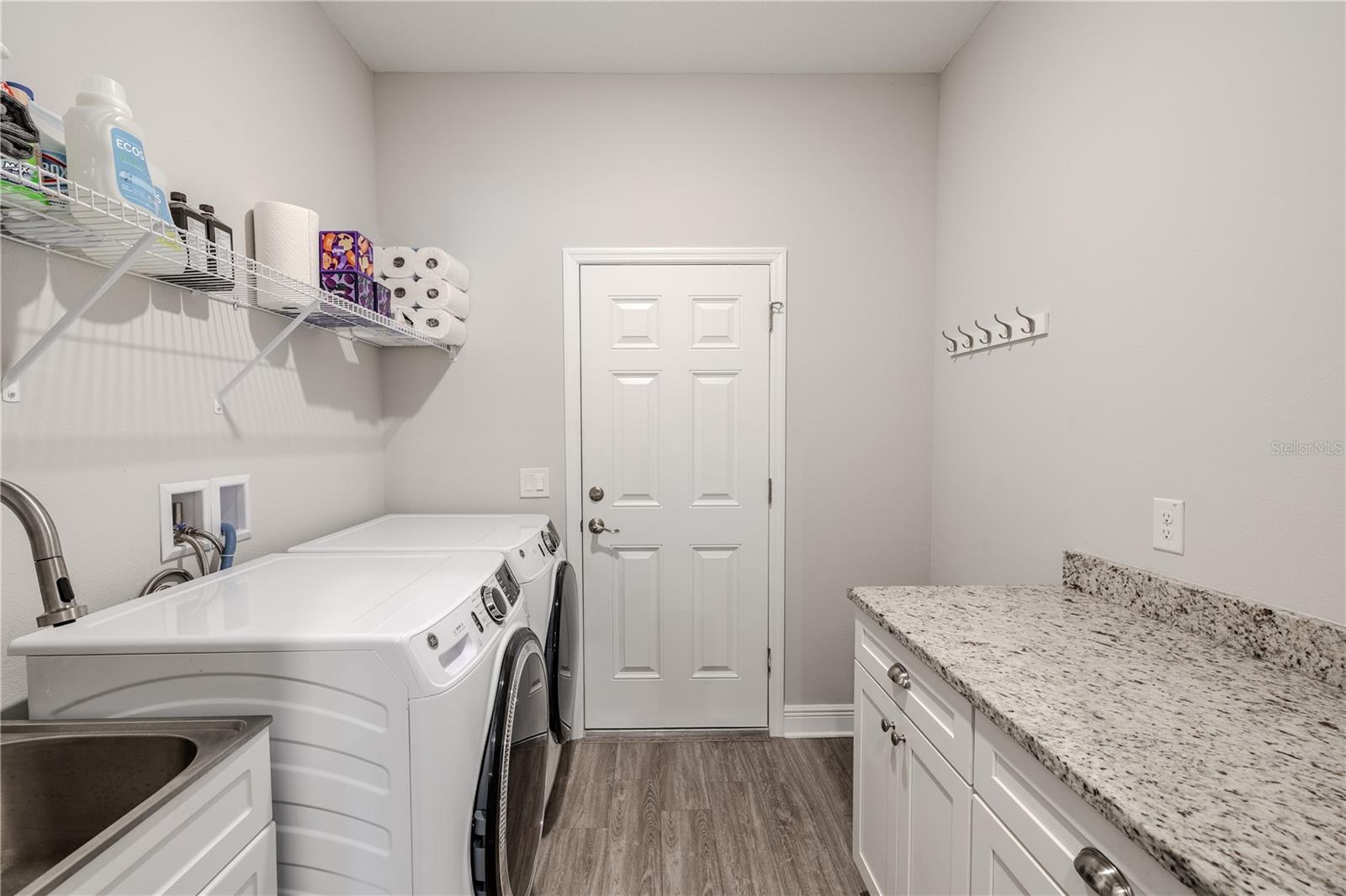 The laundry room is both functional and well-organized, featuring a side-by-side front-loading washer and dryer set beneath a wire shelf for convenient storage of laundry essentials. A stainless steel utility sink is built into the cabinetry.. Adjacent, a long granite countertop provides ample folding space, with additional cabinets below for concealed storage. The neutral wall color and wood-look flooring create a clean, modern aesthetic, while a door  leads to the garage for easy household access. A row of hooks on the wall adds a handy spot for hanging items.