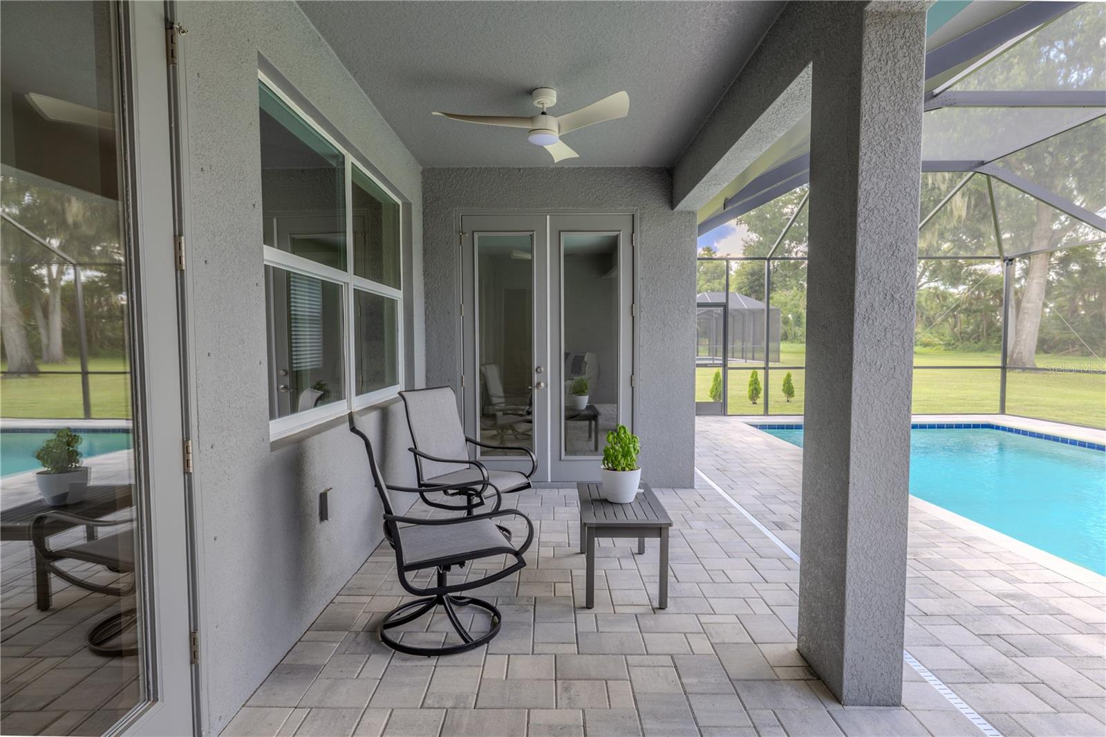The back patio features a covered, screened-in design overlooking the pool and backyard. It is finished with neutral-toned paver flooring, offering both durability and a polished appearance. A ceiling fan is installed overhead to enhance comfort, and double glass doors provide direct access to the interior of the home. The screened enclosure ensures a clear view of the pool and surrounding green space while helping keep the area comfortable and bug-free.