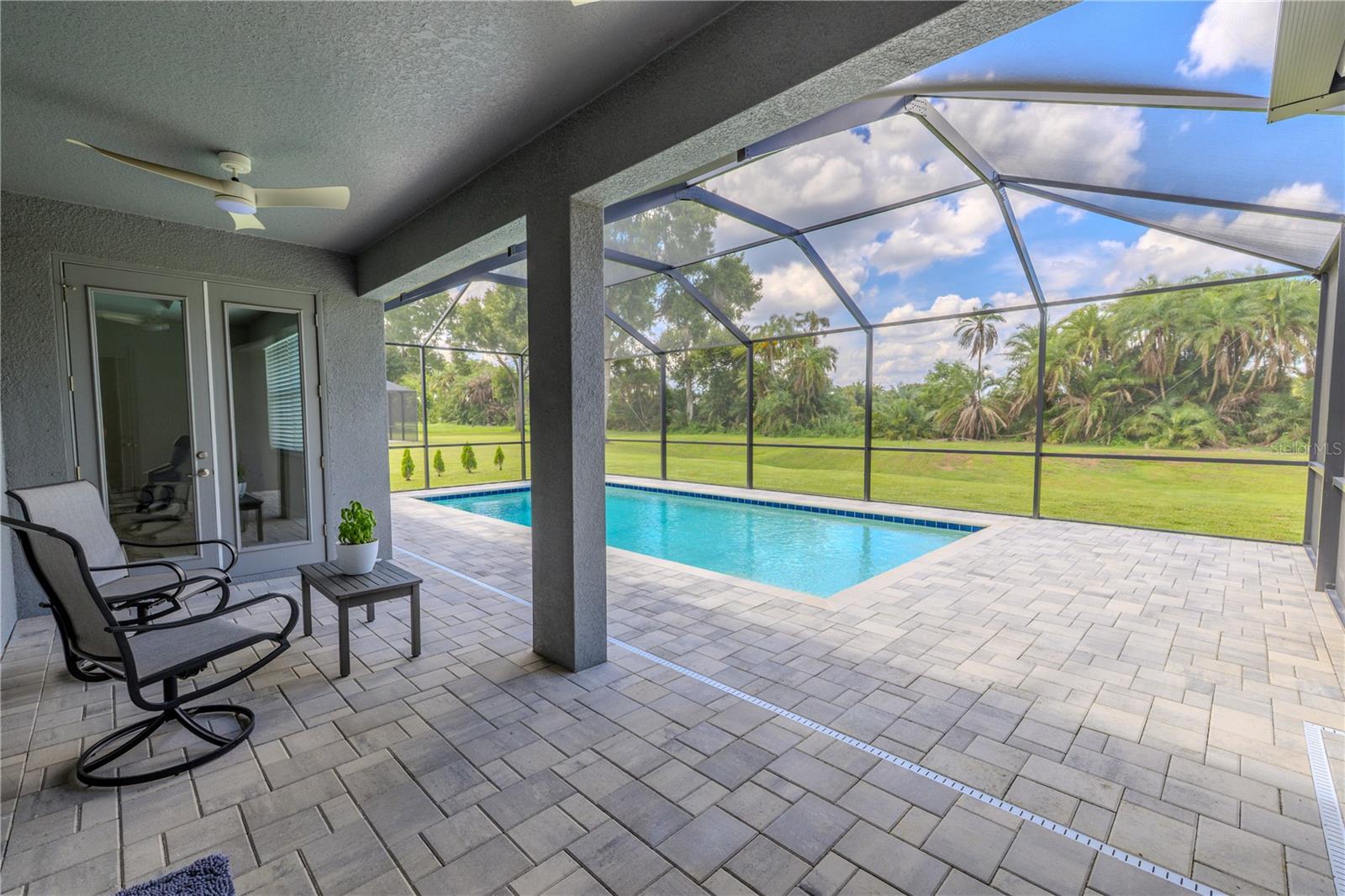 The back patio features a covered, screened-in design overlooking the pool and backyard. It is finished with neutral-toned paver flooring, offering both durability and a polished appearance. A ceiling fan is installed overhead to enhance comfort, and double glass doors provide direct access to the interior of the home. The screened enclosure ensures a clear view of the pool and surrounding green space while helping keep the area comfortable and bug-free.