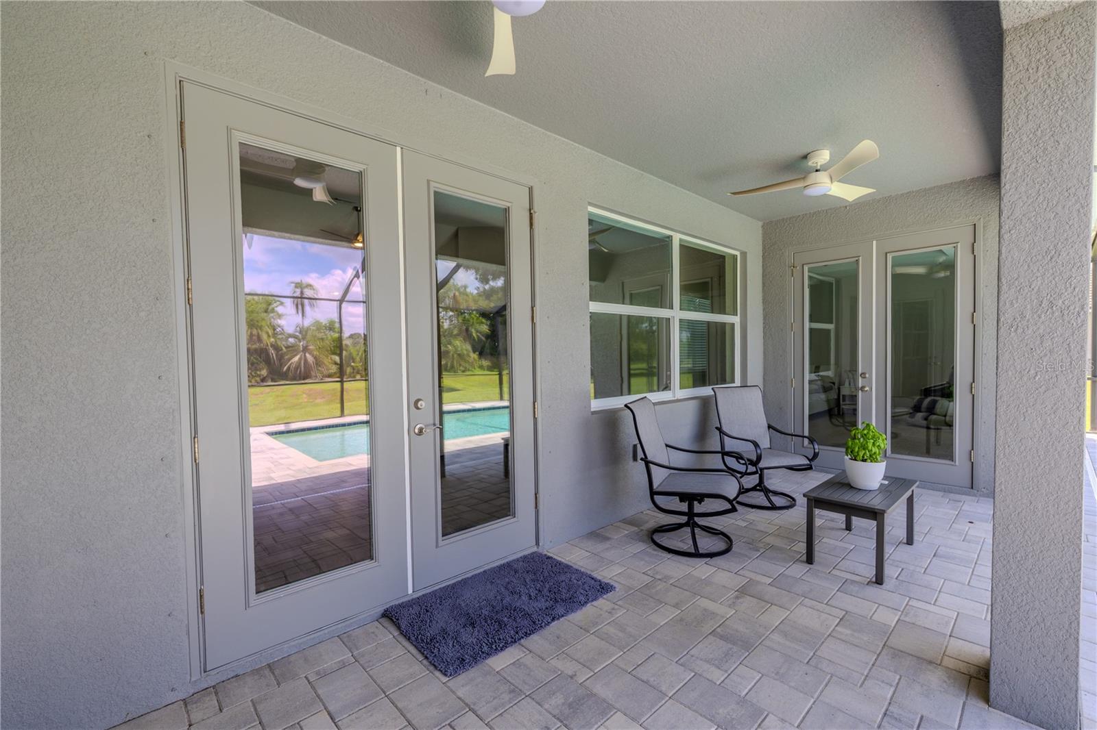 The back patios covered design with a light gray paver floor has 2 ceiling fans for added comfort. Two sets of French doors with glass panels provide access to the interior of the home, while a large window offers a clear view of the outdoor space.