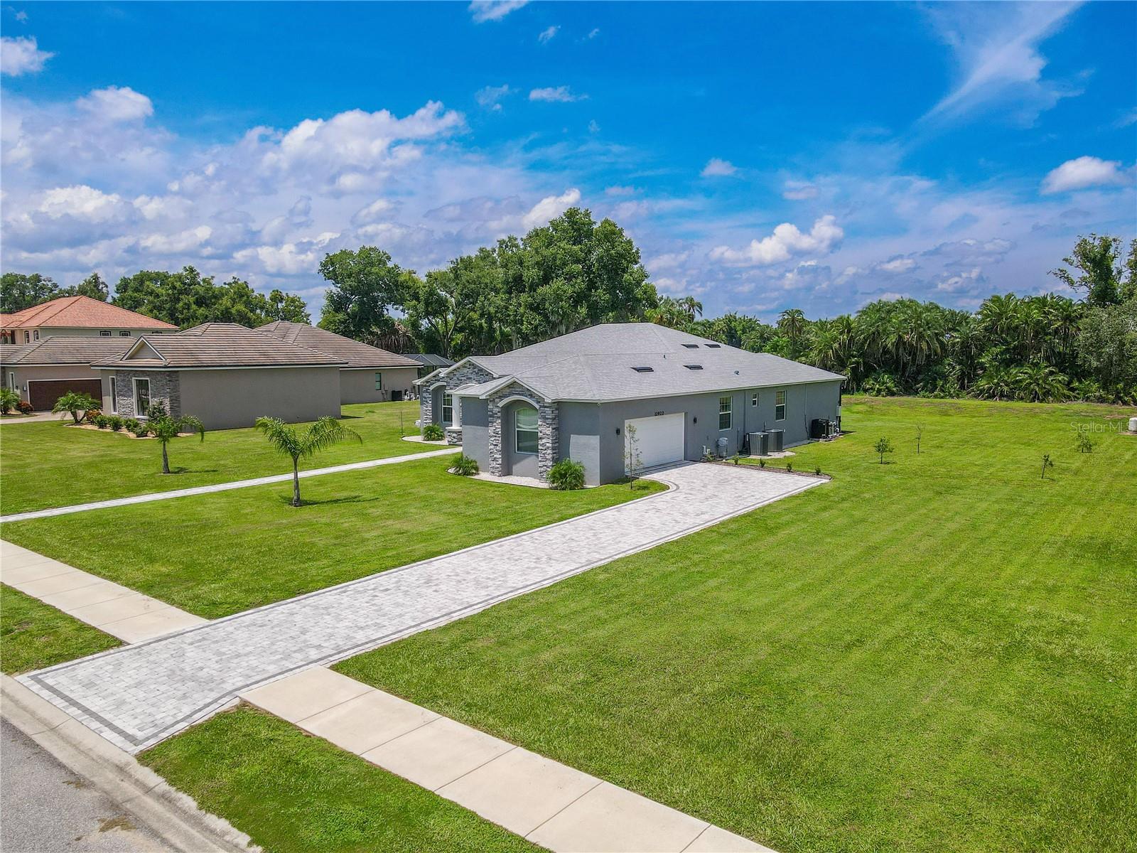 You are greeted by a light gray facade accented with stonework around the front entry. A long, paved driveway leads to a side facing, two-car garage, complemented by a matching paved walkway to the front door. The property sits on a spacious, well-manicured lawn with young landscaping and plenty of open green space on both sides. The home is positioned in a neighborhood of similar well-kept properties, with a backdrop of lush trees and palms adding privacy and a natural setting.