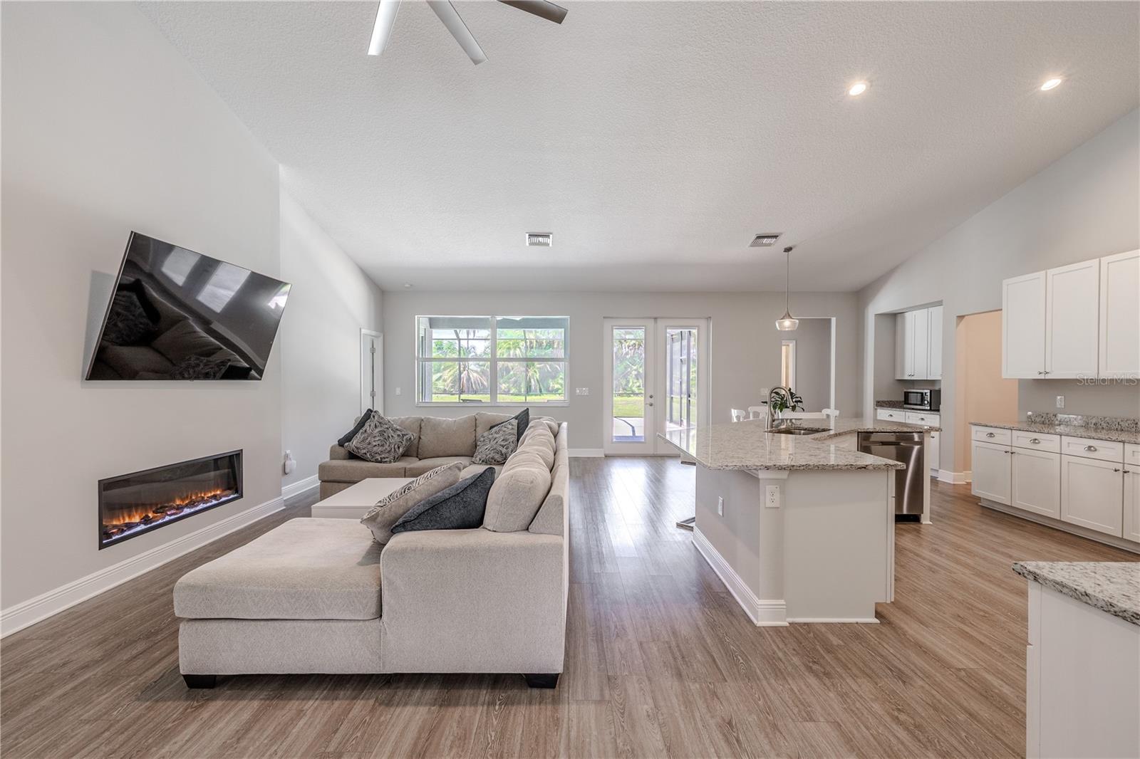 The inviting family room features a sleek electric fireplaceideal place to hang a wall-mounted TV above, creating a modern focal point for gatherings. Expansive windows and glass doors bring in abundant natural light and offer serene views of the backyard and pool area. The open layout seamlessly connects to the kitchen and dining spaces, with wood-look flooring and neutral finishes enhancing the bright, airy feel — perfect for both everyday living and entertaining.
