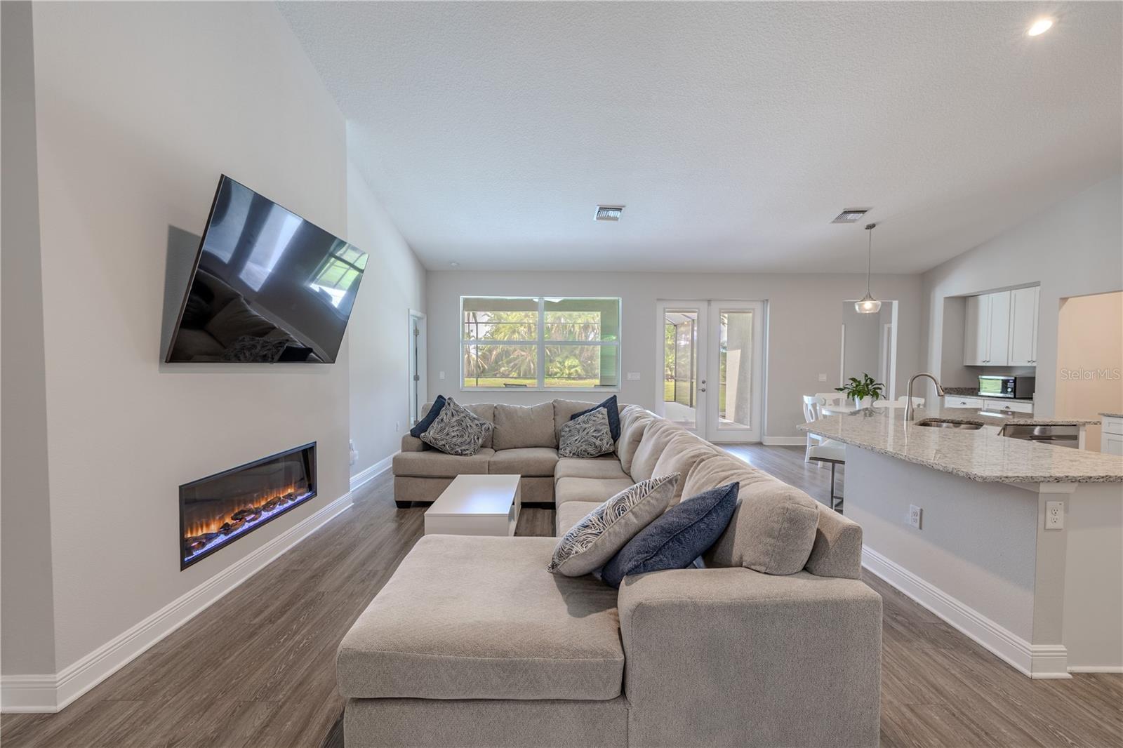 The inviting family room features a sleek electric fireplaceideal place to hang a wall-mounted TV above, creating a modern focal point for gatherings. Expansive windows and glass doors bring in abundant natural light and offer serene views of the backyard and pool area. The open layout seamlessly connects to the kitchen and dining spaces, with wood-look flooring and neutral finishes enhancing the bright, airy feel — perfect for both everyday living and entertaining.