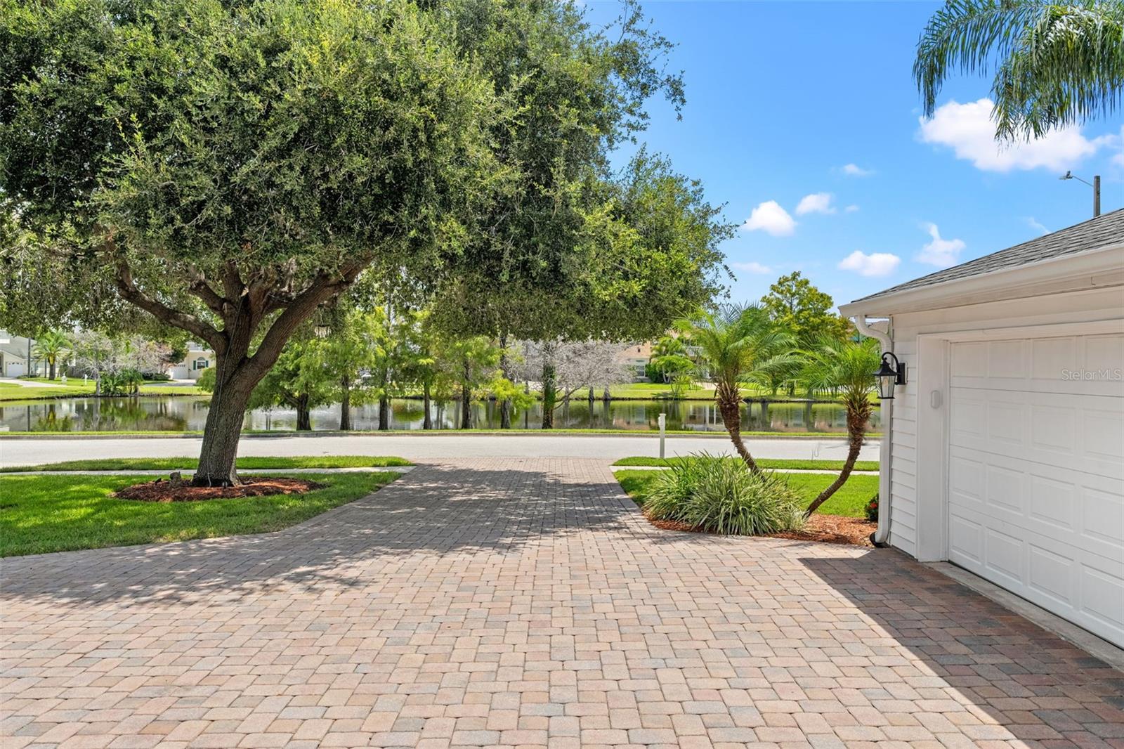 Expansive paver driveway framed by mature landscaping and serene pond views across the street.