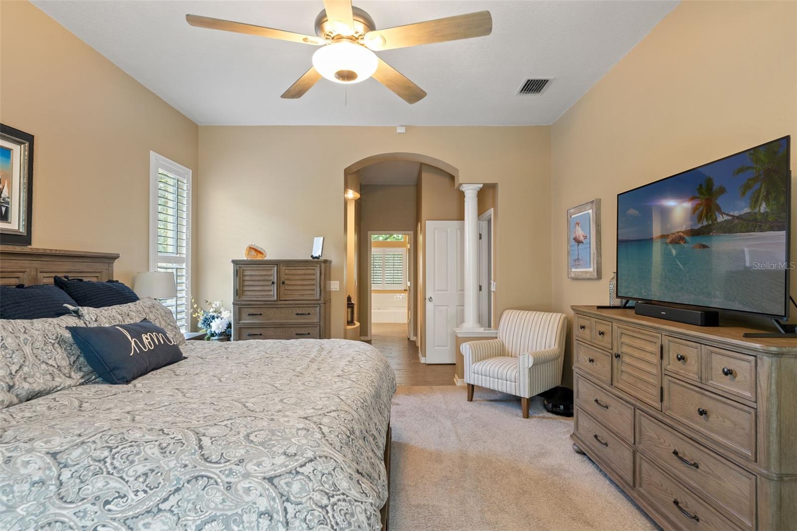 This inviting primary suite includes neutral tones, plush carpeting, and architectural details like a columned entry to the ensuite bath, creating a sense of luxury and flow.