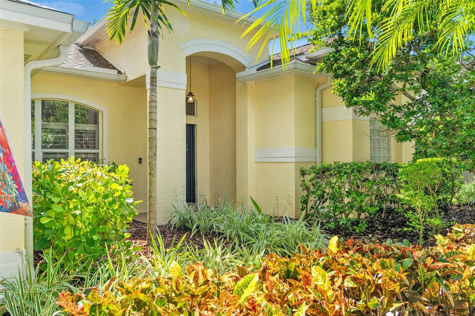 The entrance is framed by colorful plantings, palm trees, and architectural detailing. A charming front porch leads to a striking black front door with decorative glass and a transom window that welcomes natural light.
