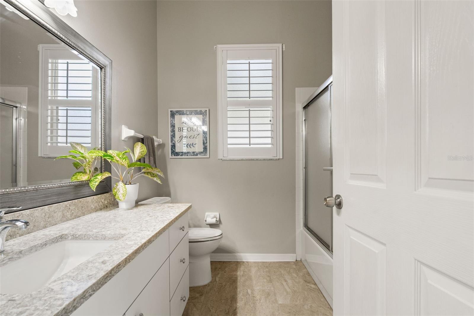 Light and bright guest bath featuring a stone-look countertop, updated vanity, and frosted glass sliding tub/shower combo beneath plantation shutters.