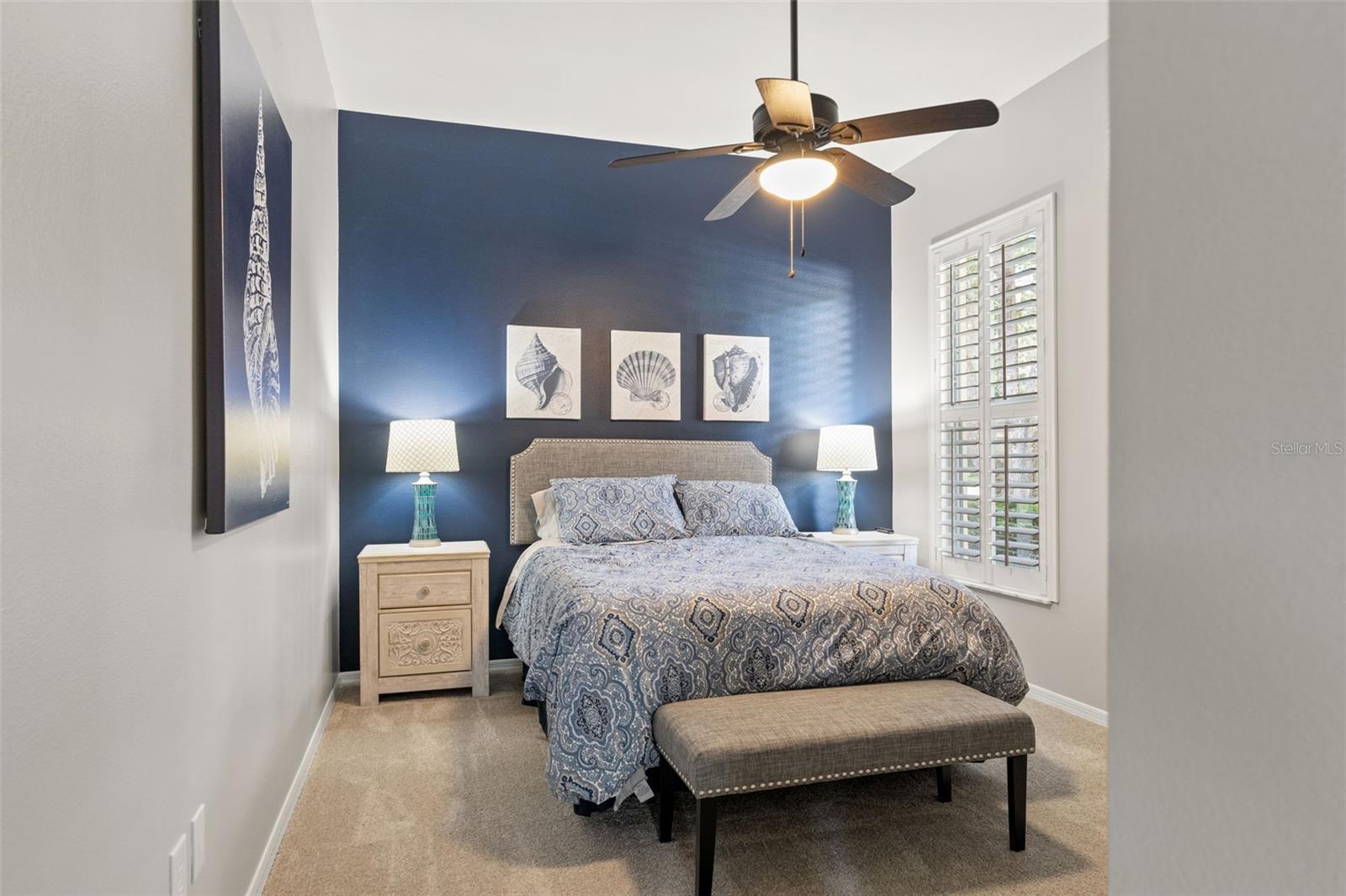 A serene guest suite with plush carpeting, coastal-inspired accent wall, and large windows with plantation shutters that fill the room with natural light.