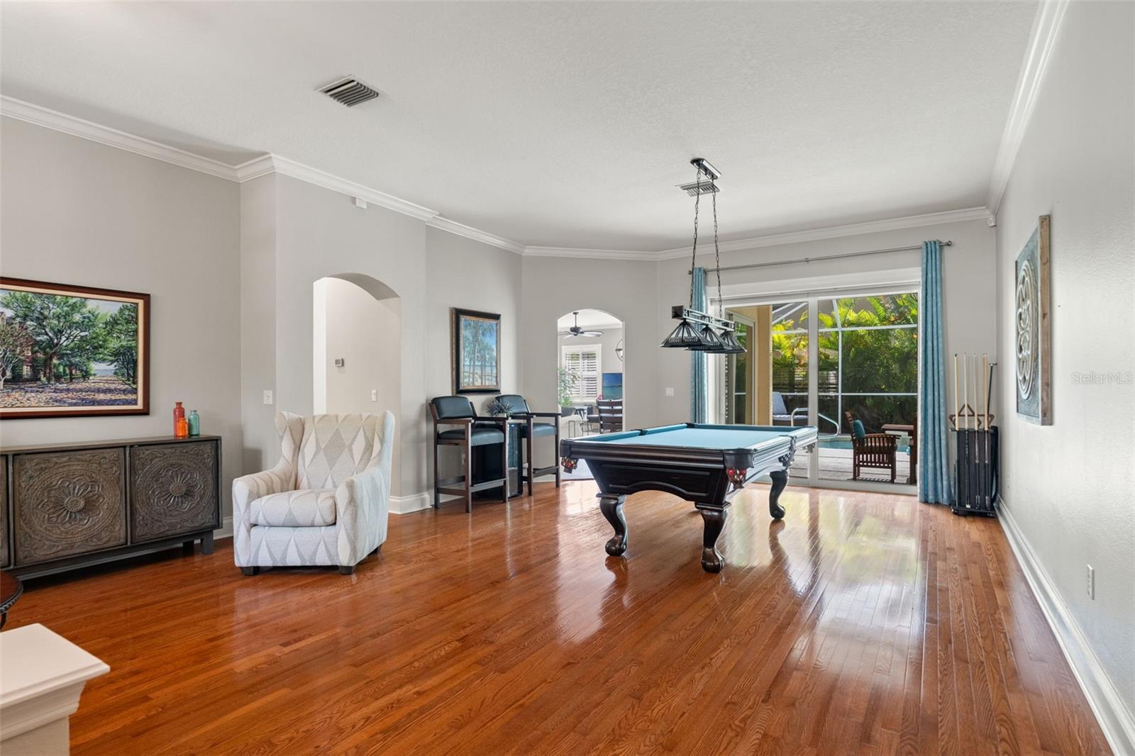 A spacious formal dining room showcased as a multi-use room featuring rich hardwood flooring, and  crown molding. Large pocket sliding glass doors open to a lush private pool/lanai area, blending indoor recreation with outdoor relaxation.