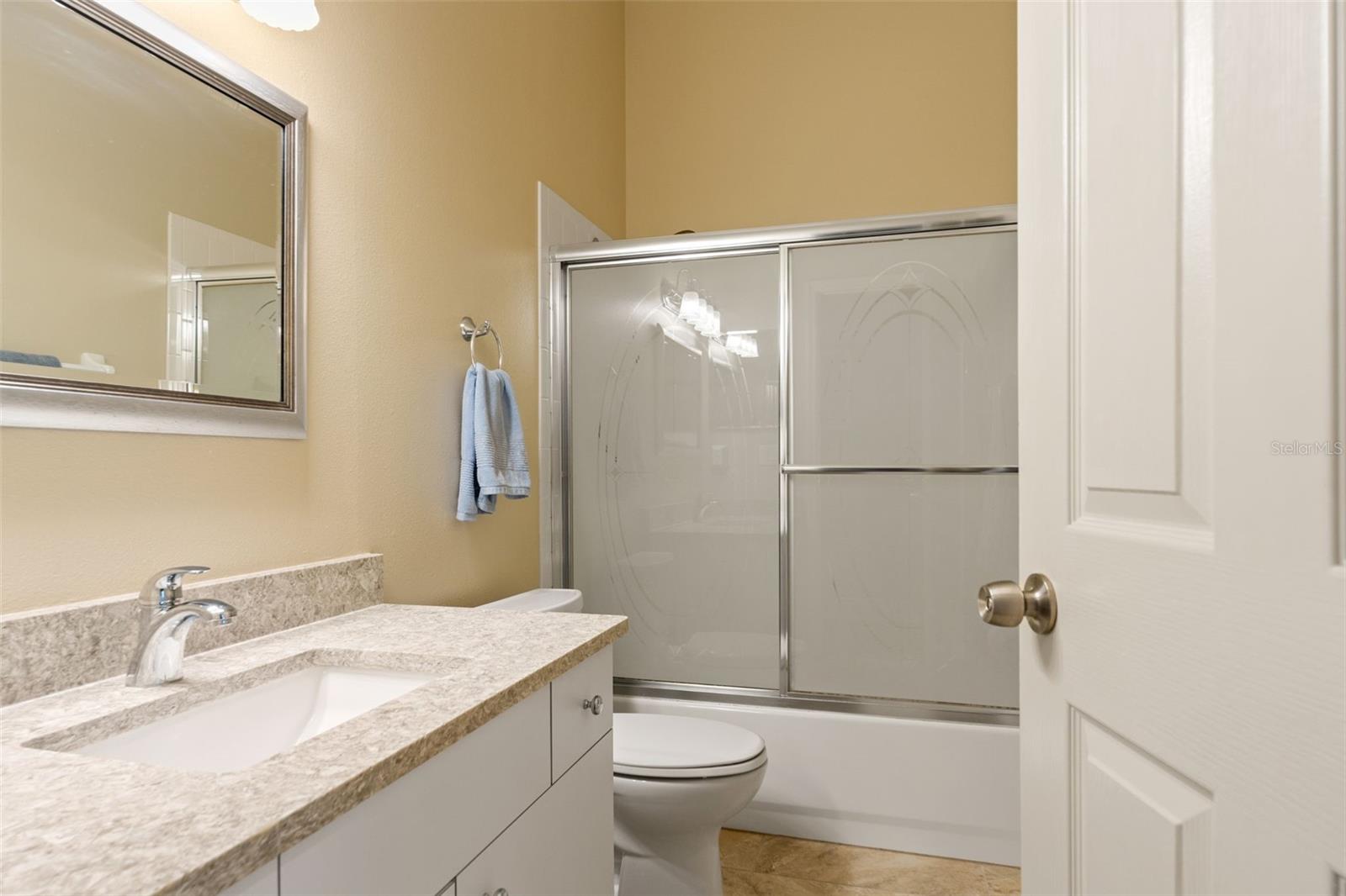 Another full bathroom with a neutral color palette, stone-look counters, and tub/shower combo with frosted sliding glass doors—convenient to both guest rooms.