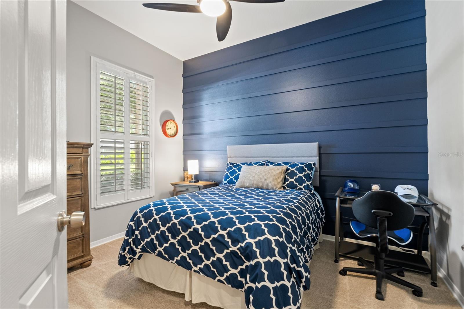 A versatile guest space with a deep navy accent wall, ceiling fan, and large window. Perfect for hosting overnight guests or creating a home office.