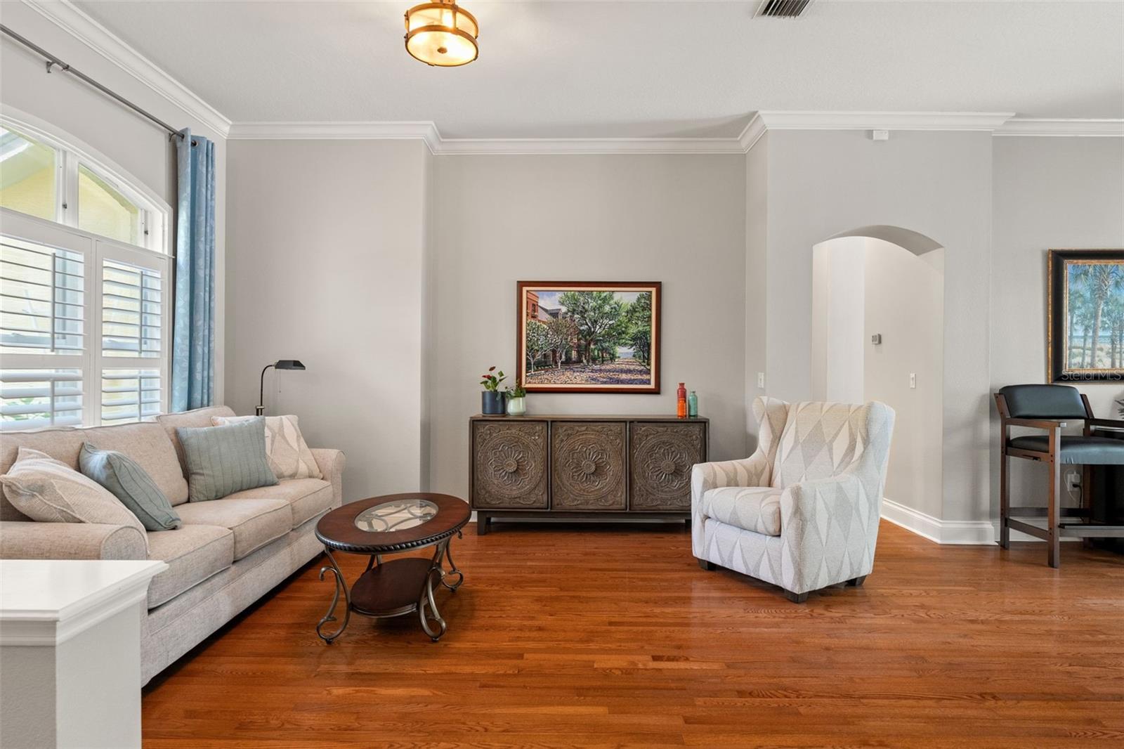 This cozy corner family room features a plush sofa, armchair, and large windows with plantation shutters—perfect for reading, relaxing, or casual conversation.