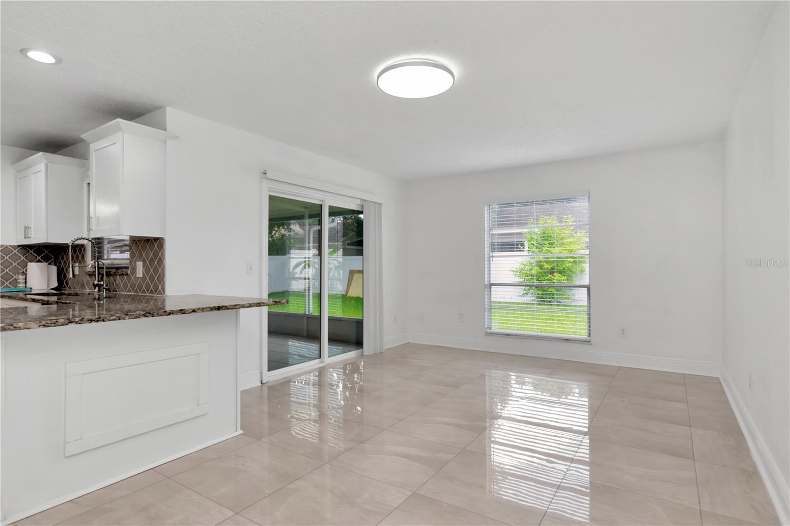 Enjoy a spacious open view from the granite counter overlooking the living and dining areas, with patio access framed by lush greenery and fruit trees.
