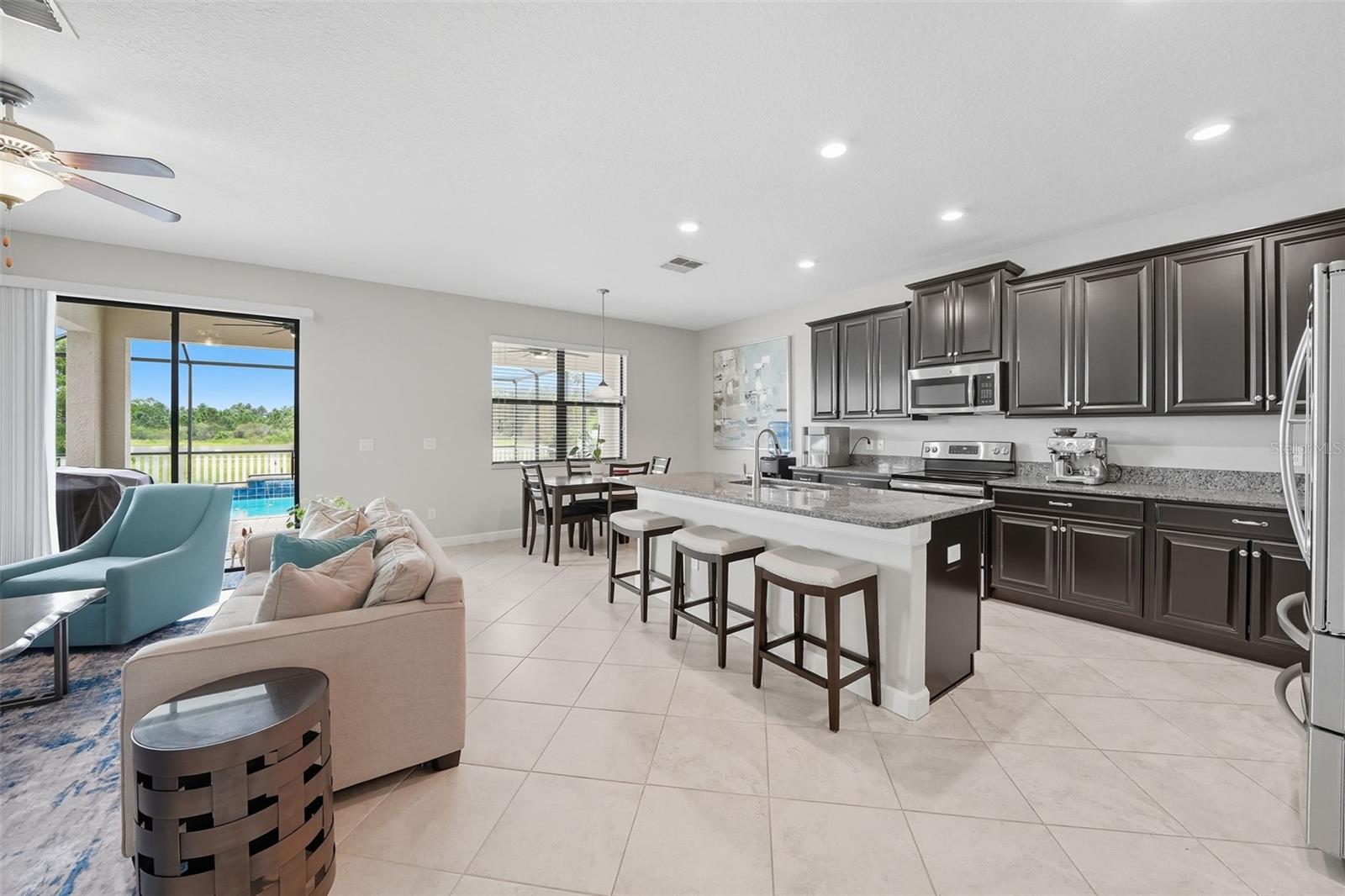 Bright open-concept living with oversized granite island and barstool seating, dining area filled with natural light, and sliding doors leading to the screened pool and patio