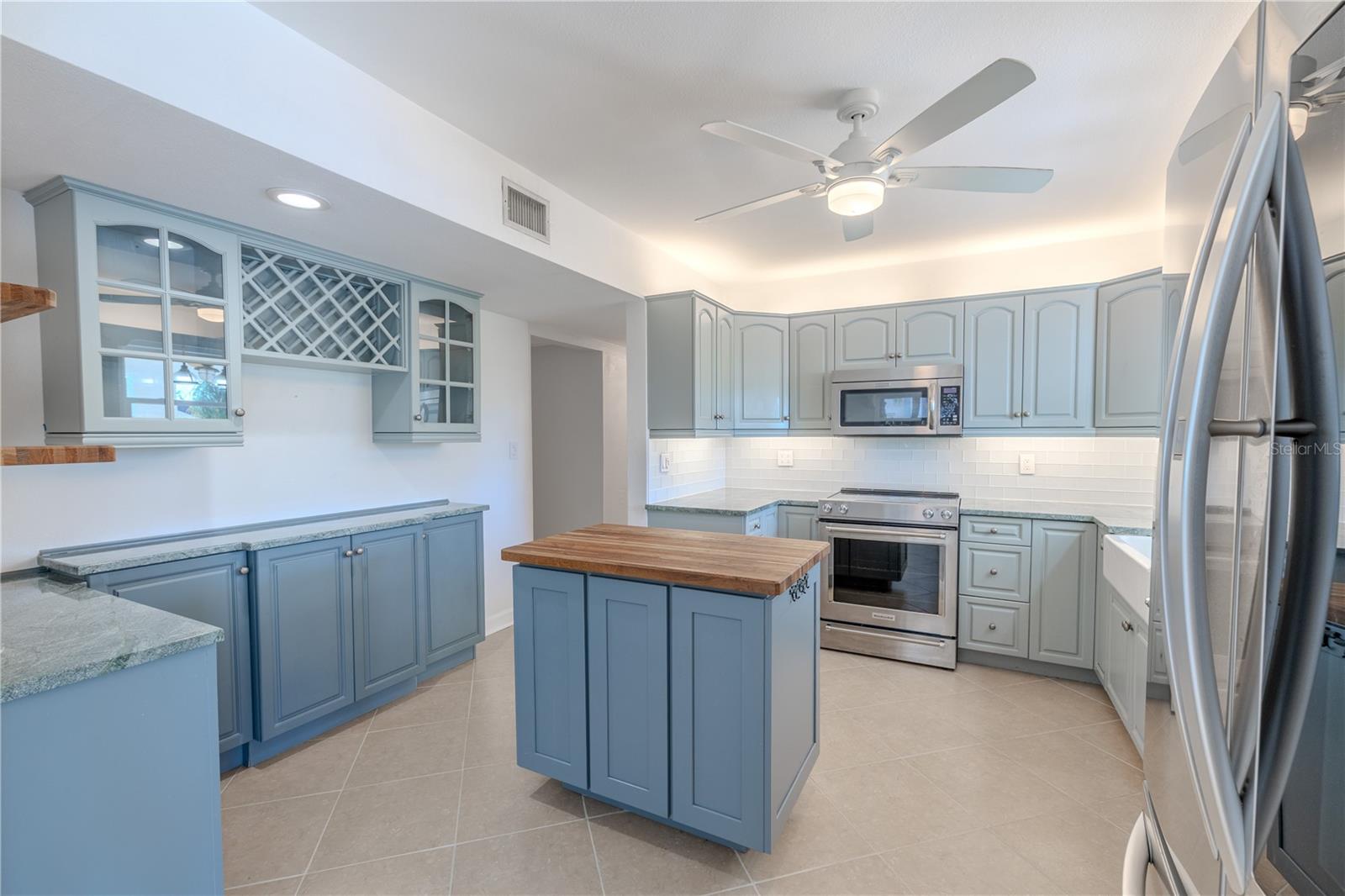 This thoughtfully designed kitchen is a blend of custom finishes featuring Mont Krest quartzite countertops, soft blue custom cabinetry, and a classic subway tile backsplash, the space offers both durability and timeless style. A central island with a butcher-block top provides additional prep and serving space, while a built-in wine rack with glass-front display cabinets adds a touch of elegance for entertaining.