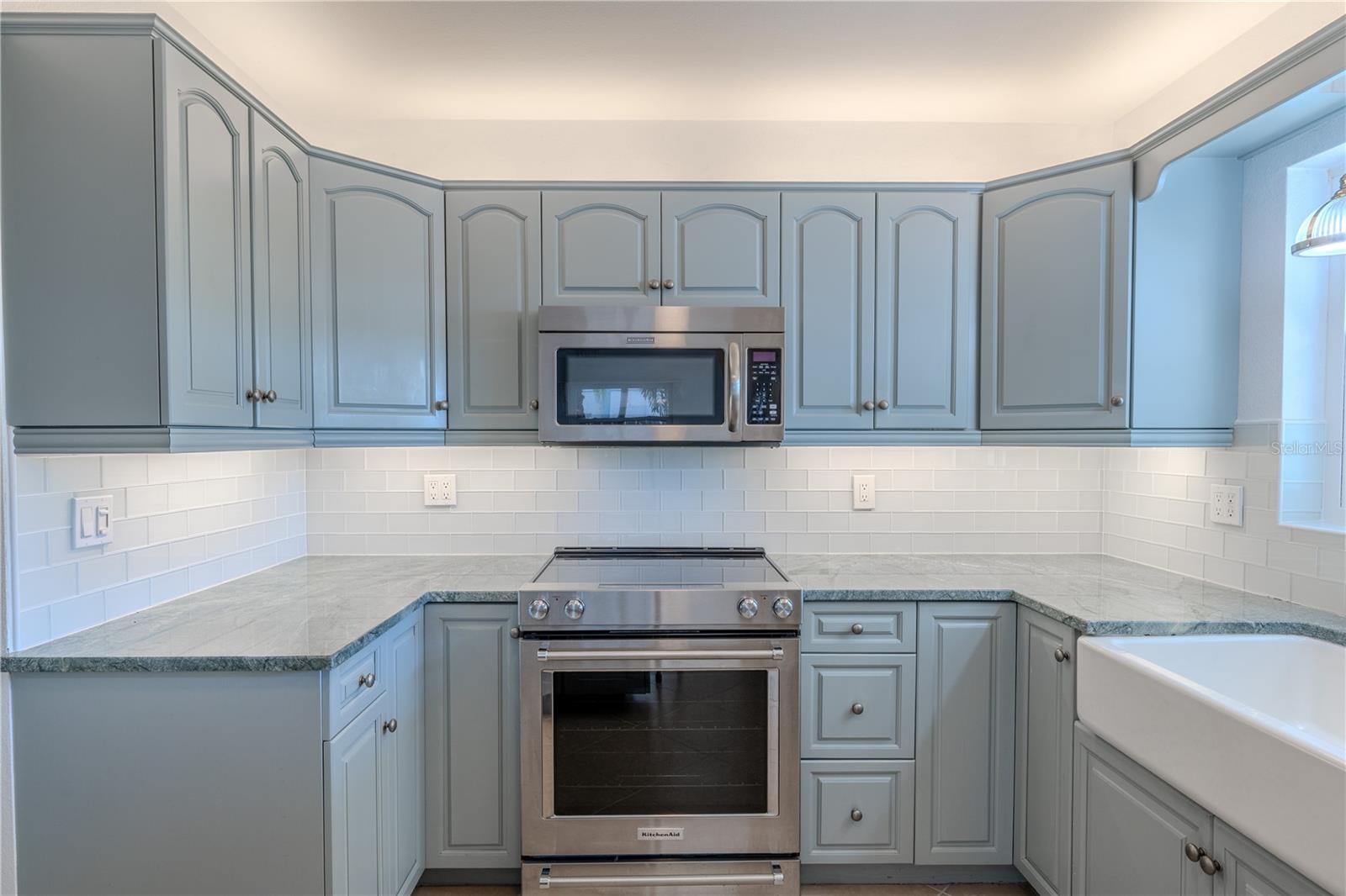 Soft blue cabinetry with classic raised-panel doors offers abundant storage, accented by brushed hardware. The stainless steel KitchenAid range and built-in microwave are framed perfectly within the cabinetry, while a farmhouse sink sits just beyond, adding charm and practicality. Under-cabinet GE dimmable lighting enhances both the countertops and backsplash, creating a bright, inviting workspace ideal for cooking and entertaining.