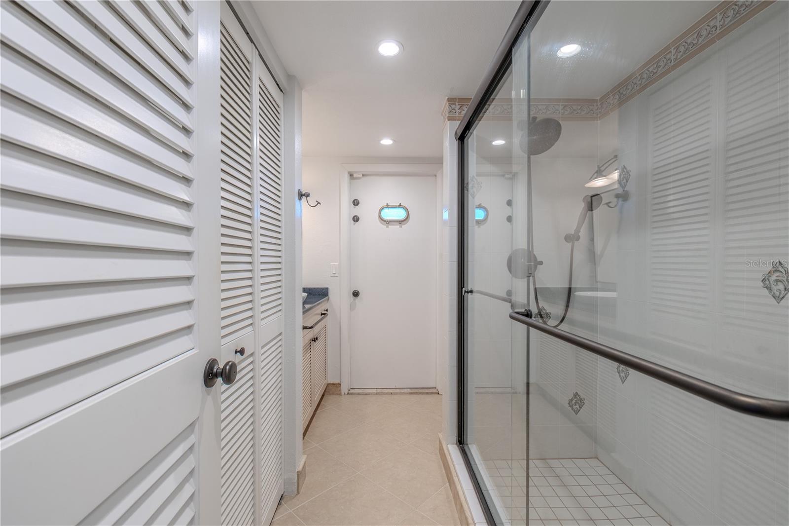 This convenient 3rd bathroom/pool bath is designed for both indoor and outdoor access, making it ideal for entertaining and waterfront living. It features a walk-in shower with glass enclosure, accented by decorative tile and a multi-function showerhead. Neutral tile flooring and recessed lighting keep the space bright. A distinctive door with a porthole-style window opens directly to the pool and patio area, allowing guests to transition easily from outdoor fun to indoor comfort.