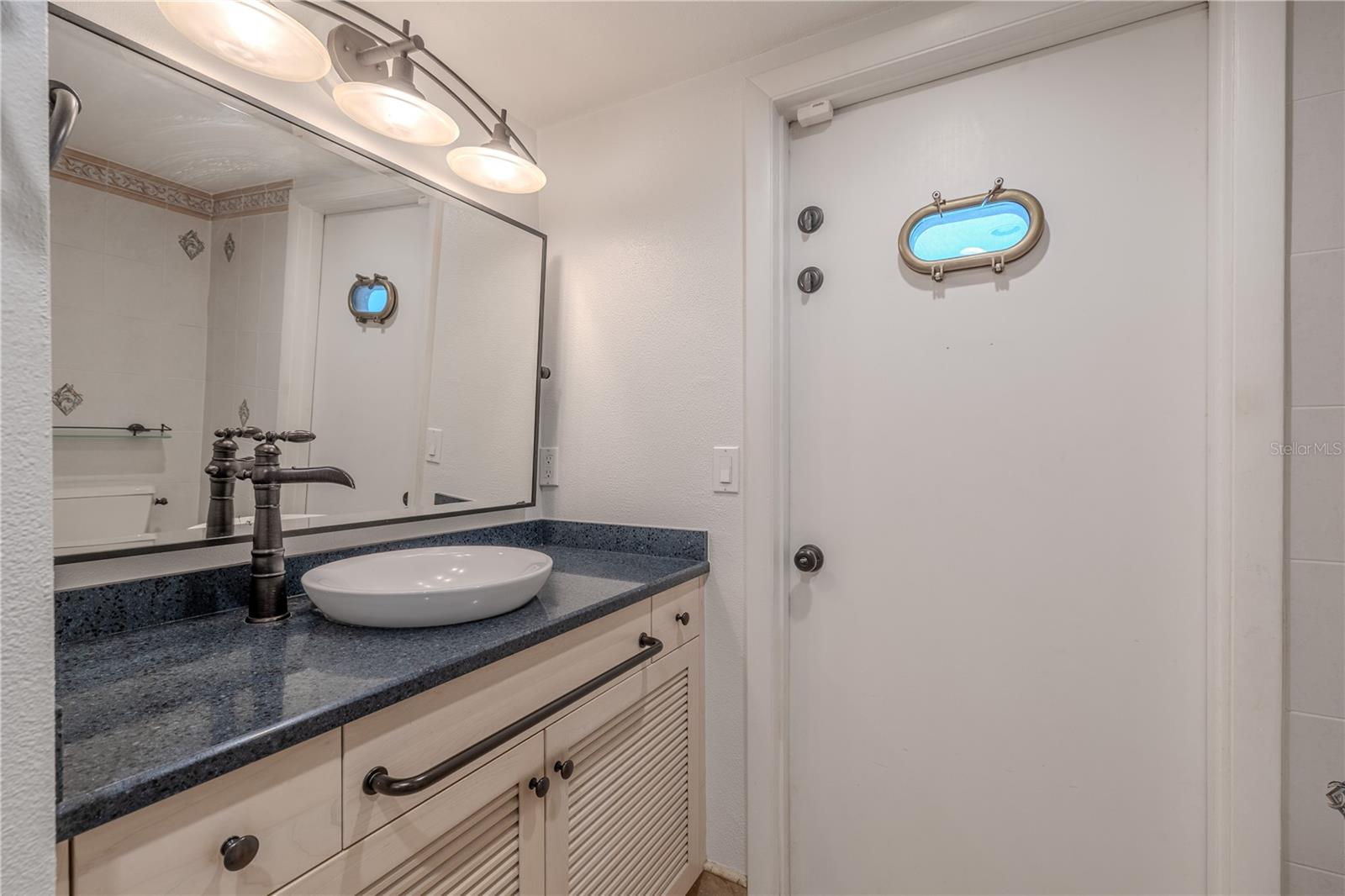 Third Bathroom – Vanity AreaThe vanity area features custom deluxe wood cabinetry with louvered doors, adding a coastal touch and extra storage. A rich solid-surface countertop is topped with a modern vessel sink, paired with Delta designer plumbing hardware in a dark finish for a stylish contrast. Overhead, a wide framed mirror and contemporary three-light fixture provide both function and elegance. This thoughtful design combines durability, character, and upscale finishes.
