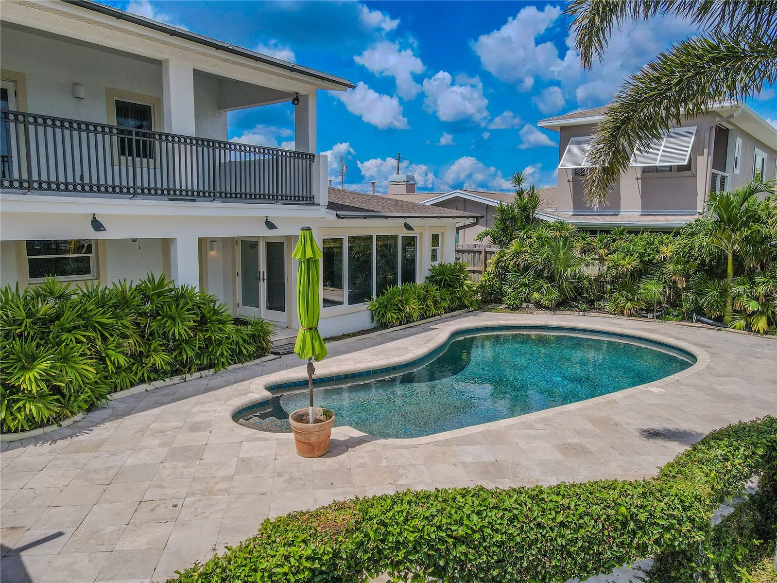 The backyard features a sparkling PebbleTec pool surrounded by an upgraded travertine deck, lush tropical landscaping, and both upper and lower covered patios for shaded relaxation.