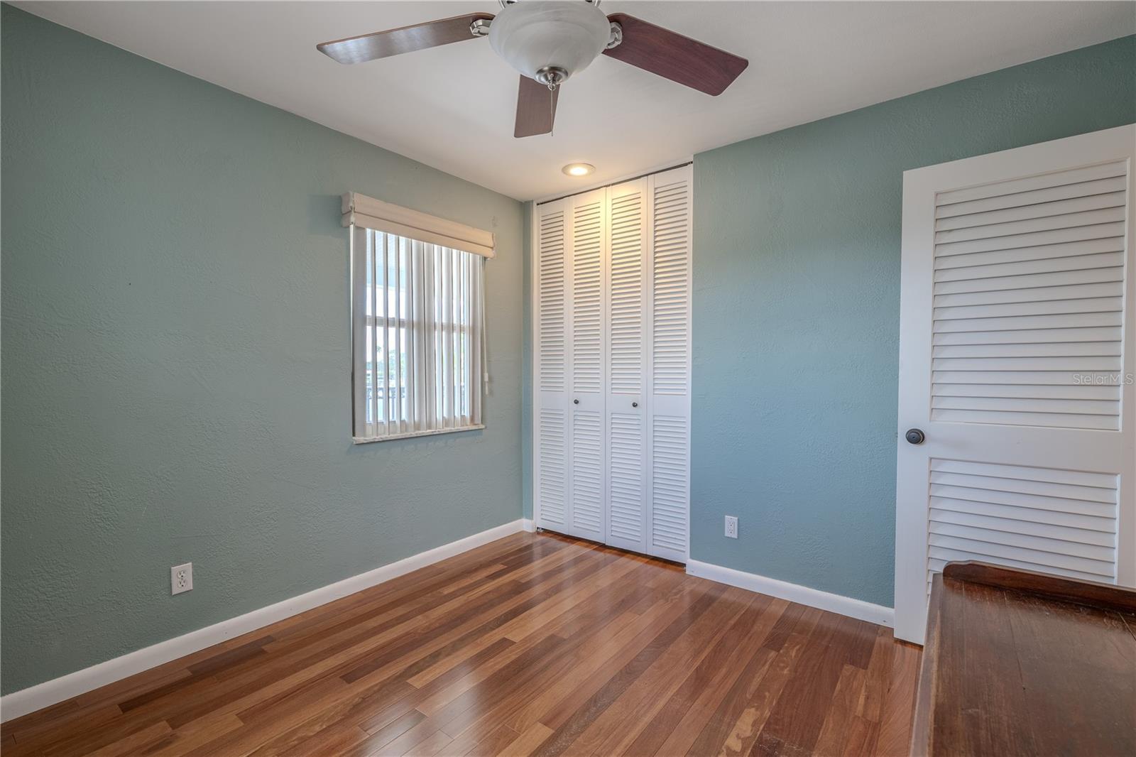 The third bedroom features Brazilian Teak solid wood flooring, a ceiling fan with light, and a built-in closet with louvered doors.