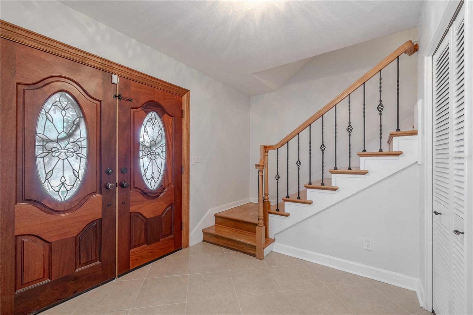 Striking custom double entry doors crafted by A1 Glass, featuring leaded glass with an elegant fleur-de-lis design that makes a stunning first impression. The foyer is finished with neutral tile flooring for a clean, timeless look, and a graceful staircase with wood steps, wrought-iron balusters, and a warm wood handrail adds character and charm.