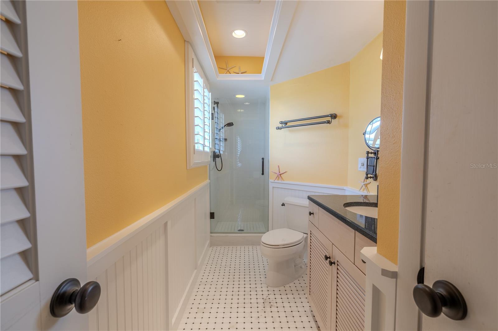 The 2nd bedroom’s ensuite bath features a marble tile floor, a Cambria quartz countertop, and custom deluxe wood cabinetry with louvered doors, blending elegance with functionality. A glass-enclosed walk-in shower with Delta designer plumbing hardware adds a touch of luxury, while custom plantation shutters provide light control and privacy. The coastal-inspired crisp white wainscoting paired with sunny walls fresh and bright.