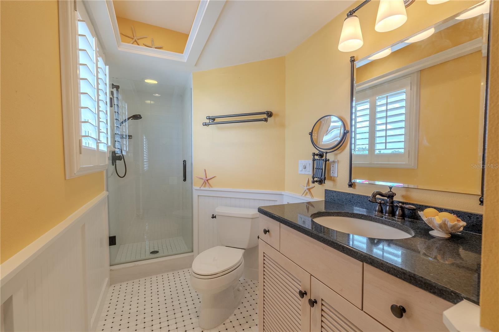 The 2nd bedroom’s ensuite bath features a marble tile floor, a Cambria quartz countertop, and custom deluxe wood cabinetry with louvered doors, blending elegance with functionality. A glass-enclosed walk-in shower with Delta designer plumbing hardware adds a touch of luxury, while custom plantation shutters provide light control and privacy. The coastal-inspired crisp white wainscoting paired with sunny walls fresh and bright.