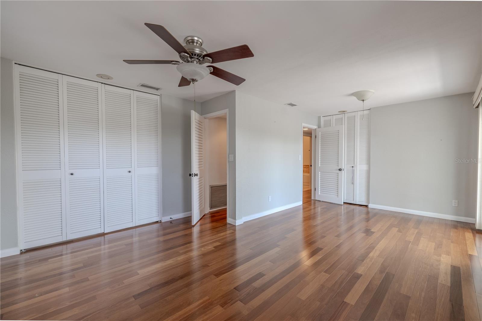 The primary bedroom has two built-in closets with louvered doors, with abundant storage and functionality. The space is finished with Brazilian Teak solid wood flooring, neutral paint, and a ceiling fan.