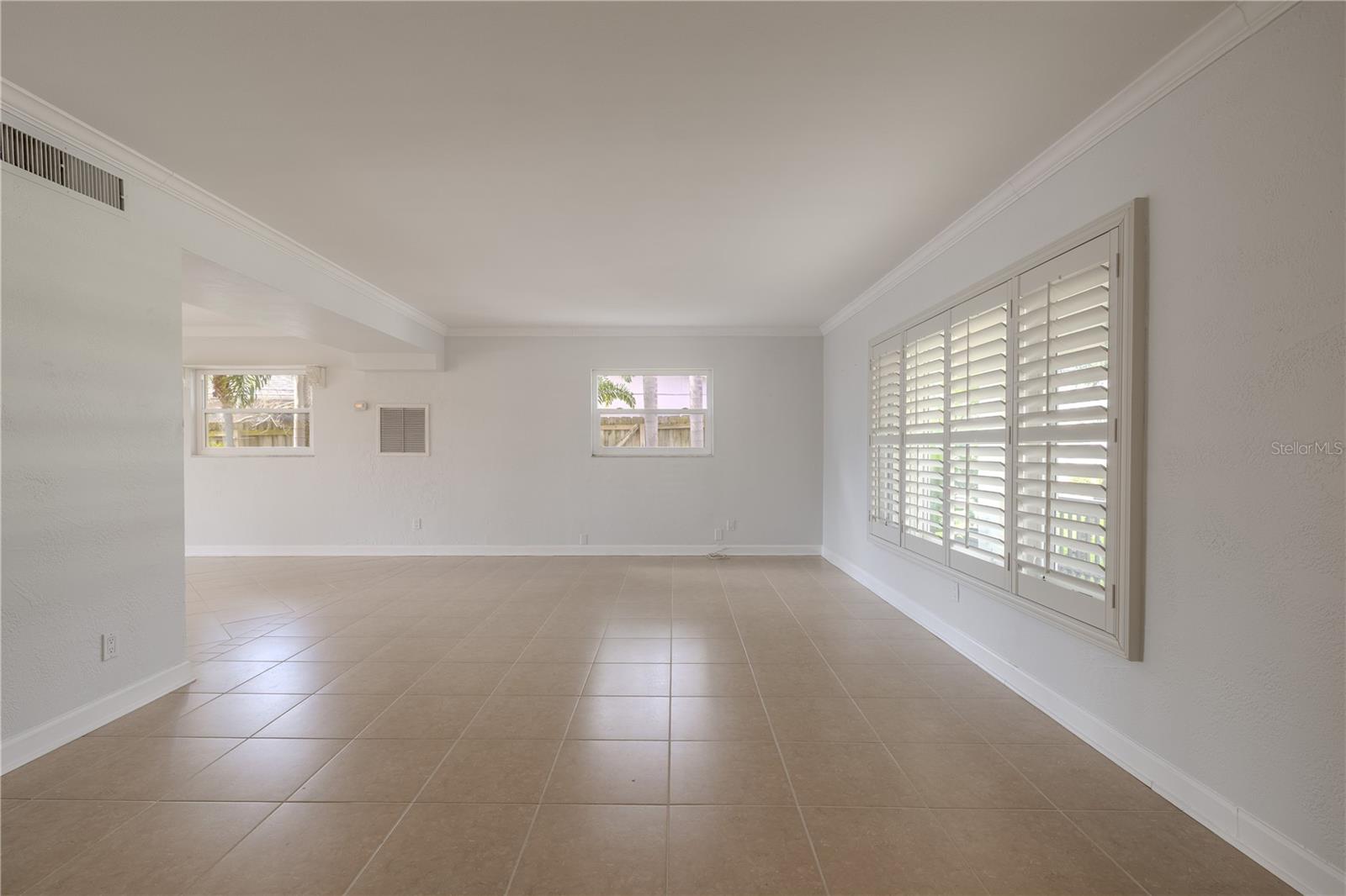 he spacious living room offers a bright and versatile layout, perfect for both relaxing and entertaining. Large windows are dressed with custom plantation shutters, allowing you to control natural light and privacy while adding a touch of timeless style. Neutral tile flooring flows throughout, complemented by soft wall tones and crisp crown molding that create a clean, sophisticated look.