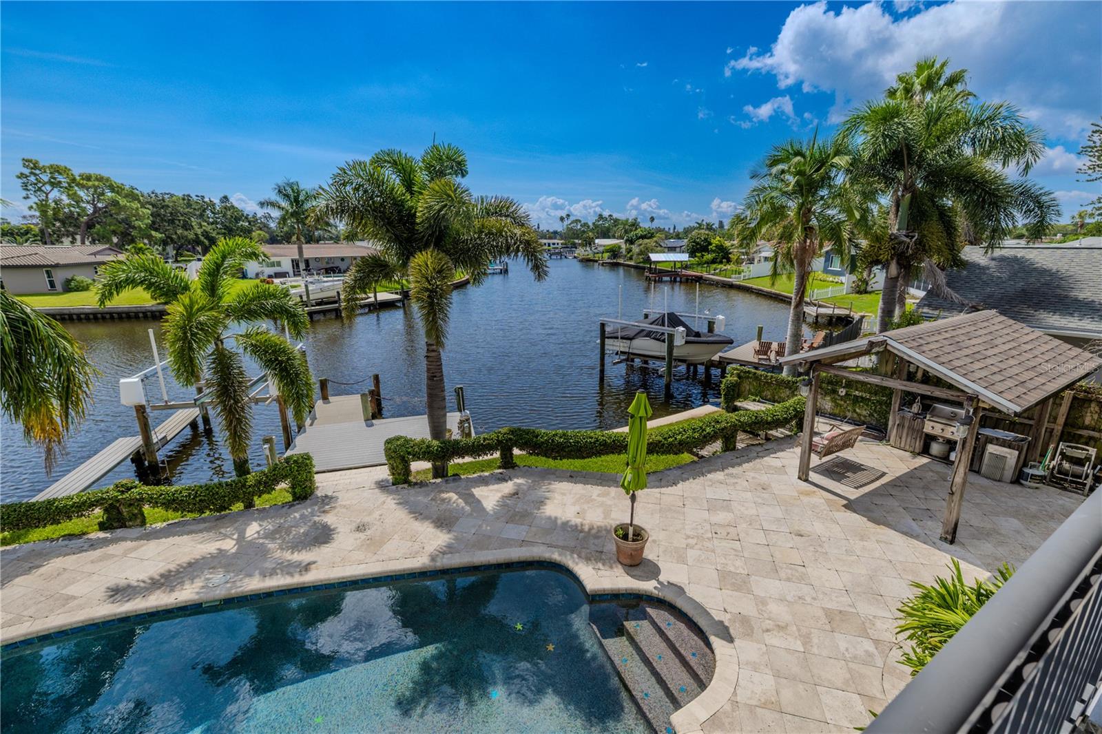 The shimmering Pebble Tec pool sparkles against its upgraded travertine deck, framed by tropical palms and lush greenery. Beyond the pool, the private dock with boat lift extends into the wide canal, offering a serene backdrop of open water.