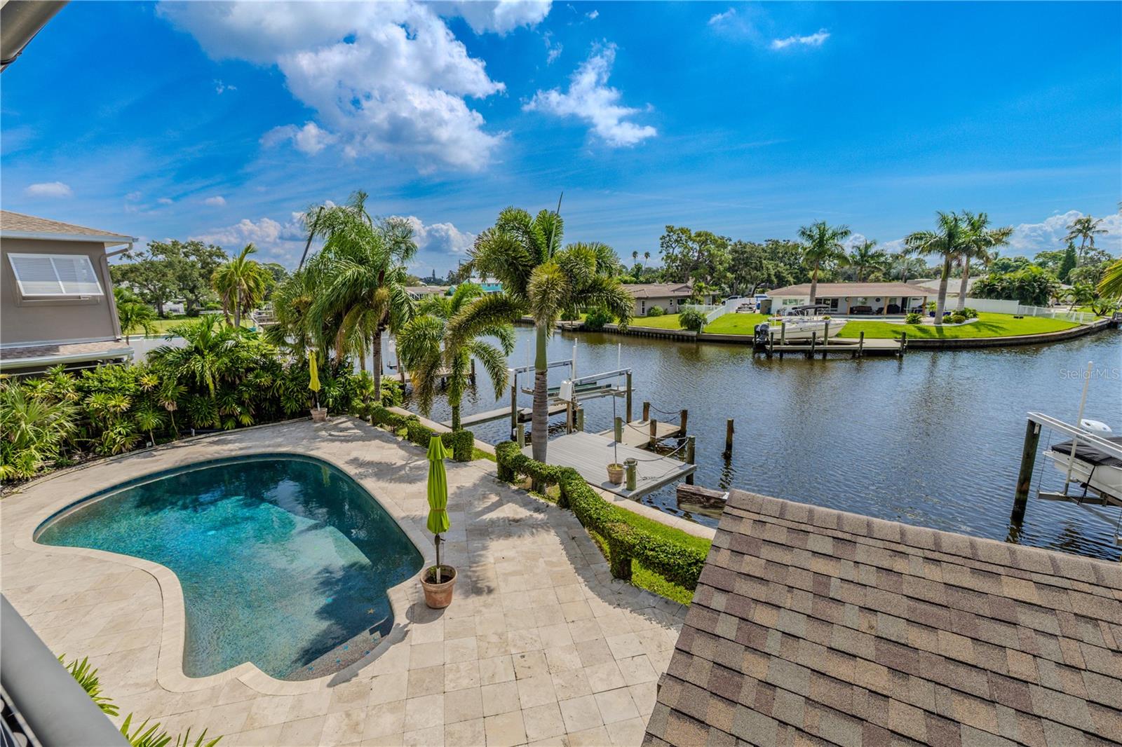 The shimmering Pebble Tec pool sparkles against its upgraded travertine deck, framed by tropical palms and lush greenery. Beyond the pool, the private dock with boat lift extends into the wide canal, offering a serene backdrop of open water