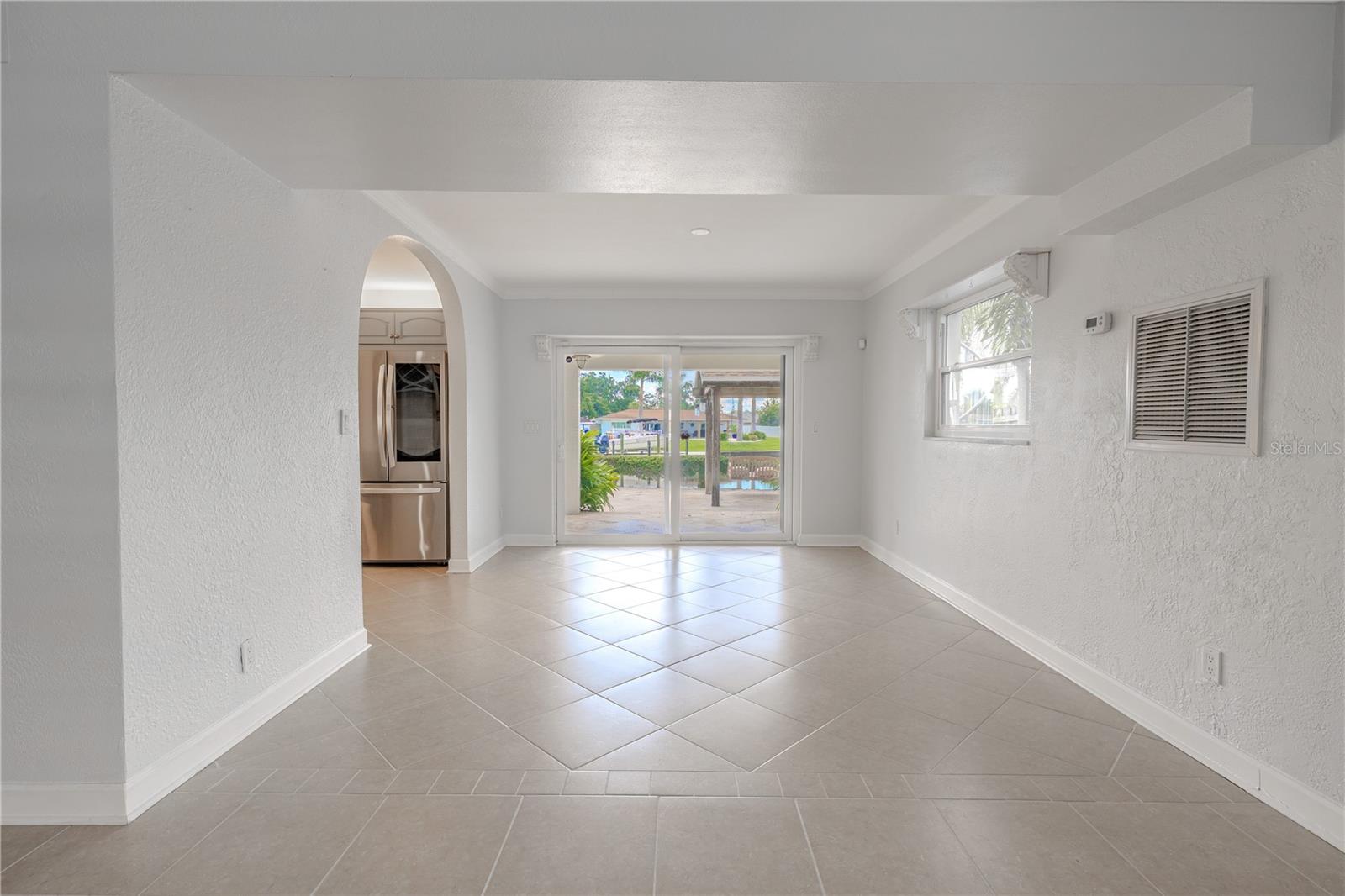 The dining room is perfectly positioned, with a bright and open layout that connects seamlessly to the kitchen and outdoor living spaces. An arched entryway leads into the kitchen, adding architectural charm, while sliding glass doors frame views of the pool, patio, and canal, creating a natural extension of the dining space outdoors. A side window adds additional natural light, and the neutral tile flooring with decorative inlay detail provides a subtle touch of elegance. This space captures the essence of indoor-outdoor Florida living.