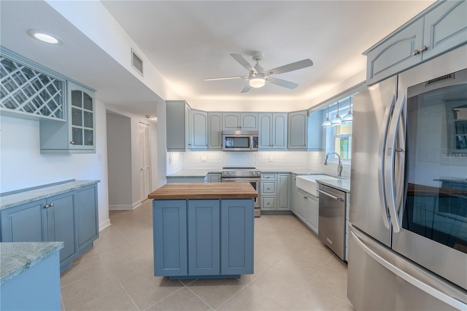 This beautifully updated kitchen blends style and functionality, featuring soft blue cabinetry with custom trim, Mont Krest custom Quartzite countertops, and a butcher-block island that doubles as prep and serving space. A farmhouse sink sits beneath a window with RH pendant lighting, while GE under-cabinet dimmable lighting enhances both ambiance and task efficiency.