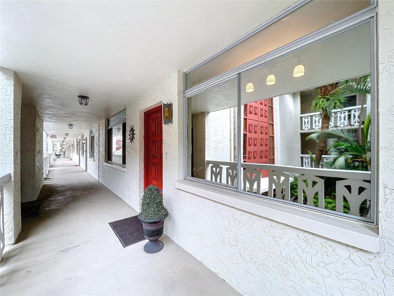 The unit is conveniently located along a well-lit walkway, with charming architectural railings and tropical greenery visible through large windows.