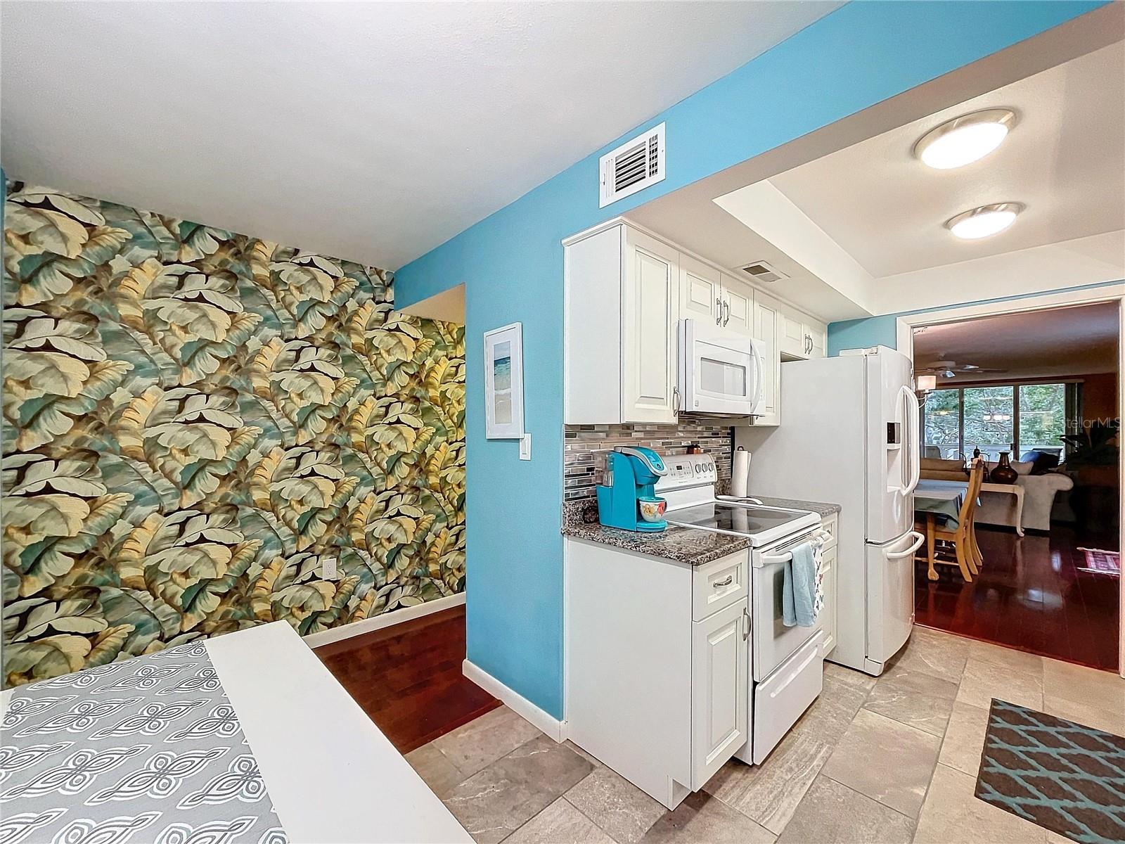 The kitchen opens directly from the dining area, featuring bold tropical wallpaper, vibrant blue accents, and convenient layout for everyday use.