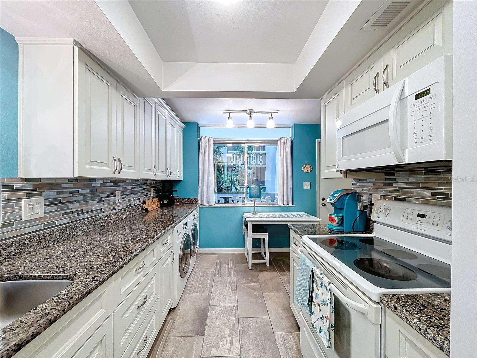 Updated galley-style kitchen with white cabinetry, granite countertops, and stainless tile backsplash—offering ample storage and prep space.
