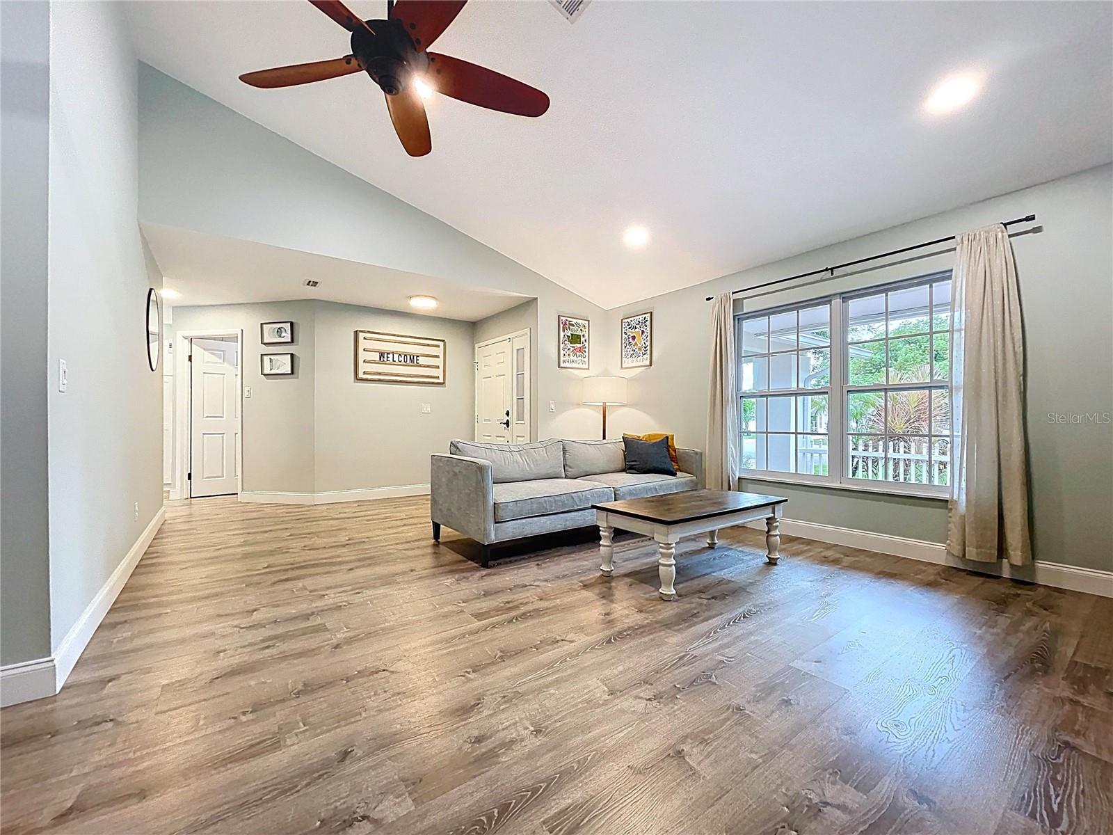 pacious and inviting living area with vaulted ceilings, wide-plank LVP flooring, and abundant natural light from a large picture window.