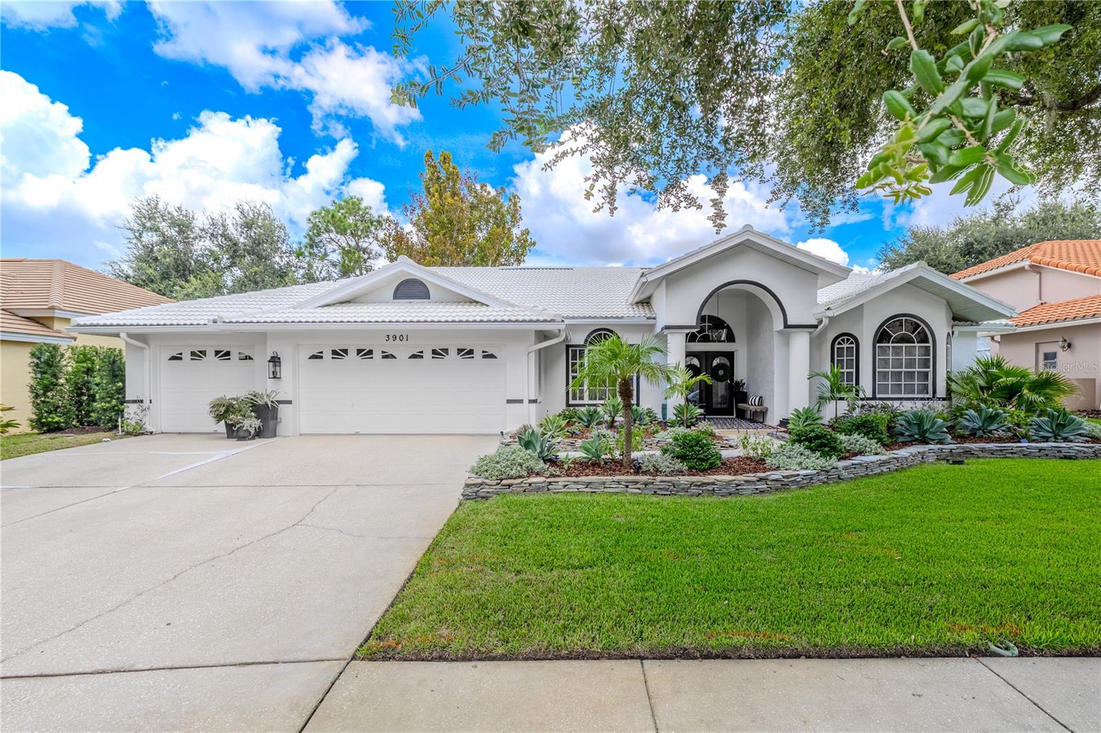 Front exterior view of the home featuring a three-car garage, wide driveway, covered entryway with arched design, and landscaped yard with stone borders and mature plantings.