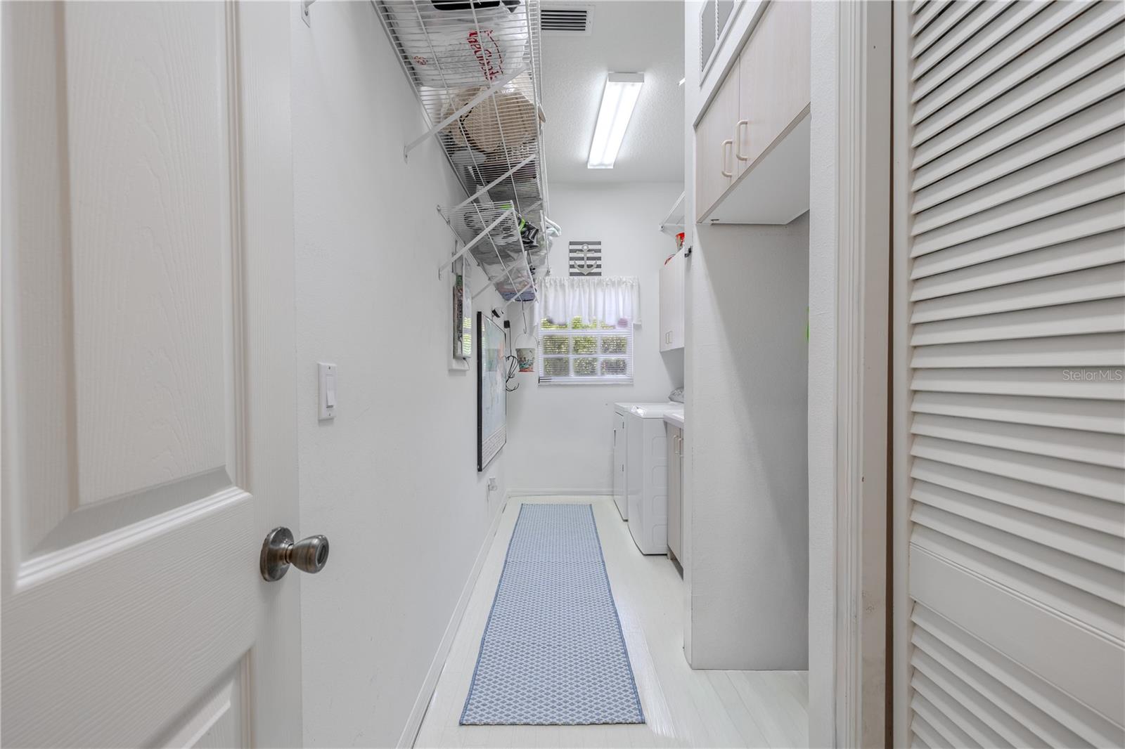 Bright laundry room with upper and lower cabinetry, overhead shelving, and a window for natural light.
