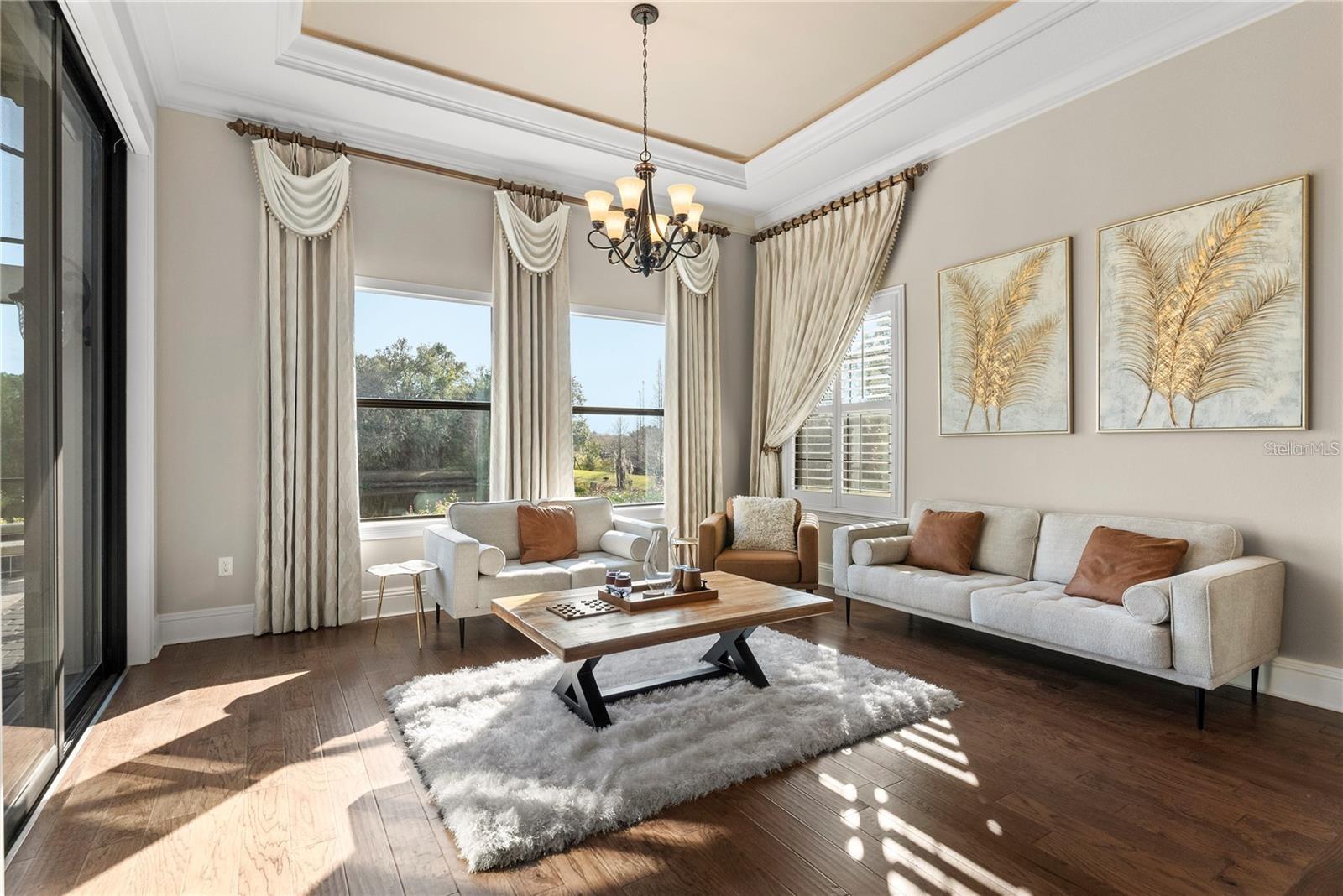 The primary bedroom’s sitting area offers a tranquil retreat, featuring rich wood floors, softly illuminated tray ceilings, and serene views of the picturesque pond.