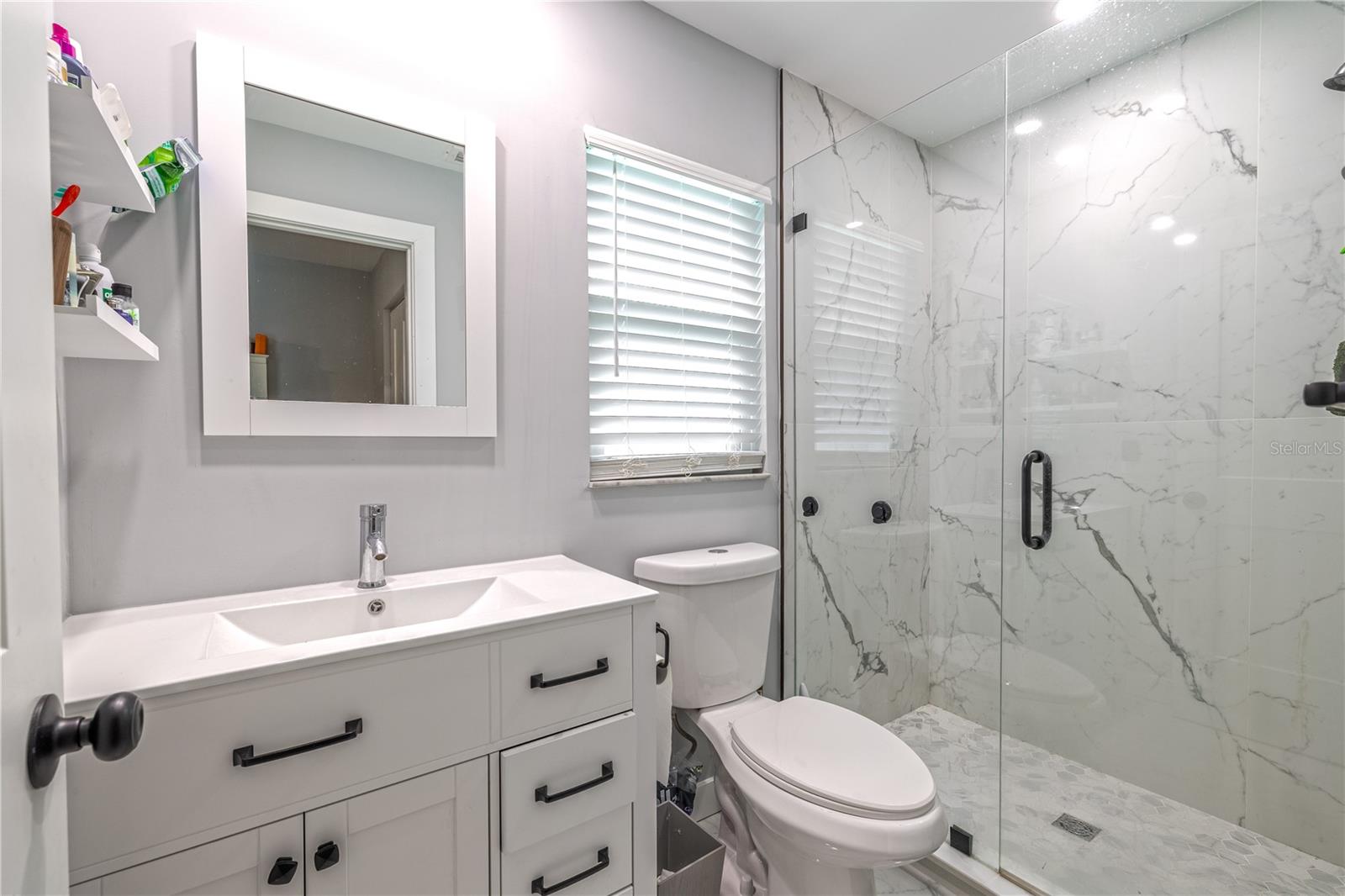 Primary bathroom featuring a frameless glass walk-in shower with marble-look porcelain tile, single vanity, and updated matte black hardware.