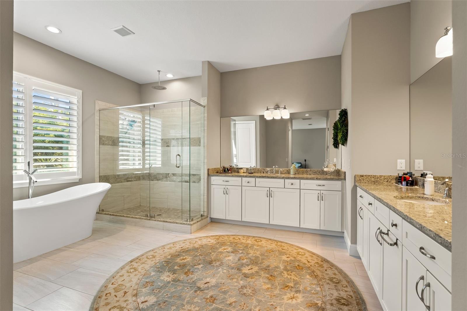 Spa-inspired bathroom featuring dual granite vanities, a soaking tub, frameless glass shower, and plantation shutter window.