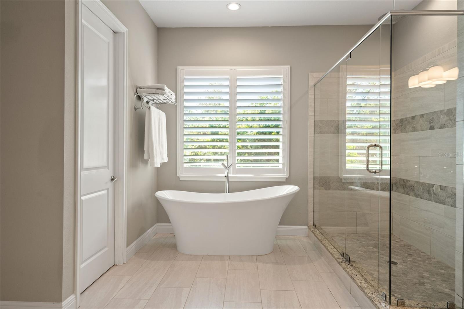 Freestanding tub positioned beneath plantation-shuttered window, paired with a sleek floor-mounted faucet.