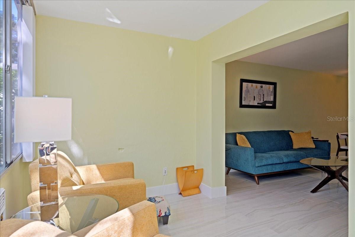 The spacious living room includes a floating entertainment center and flows seamlessly into the finished lanai/sunroom, where large windows stretch across the back of the unit, filling the space with natural light and framing tranquil views of mature oak trees and greenspace.