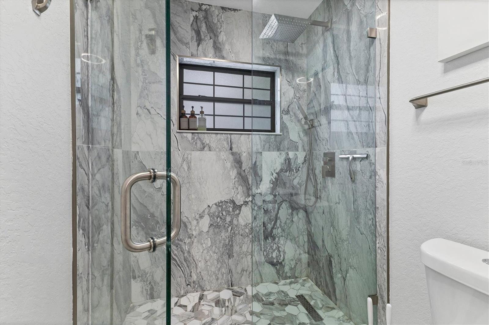 Beautifully remodeled bath with walk-in shower featuring large marble tiles, geometric tile floor with slim drain and a dual rain shower head with hand-held option.