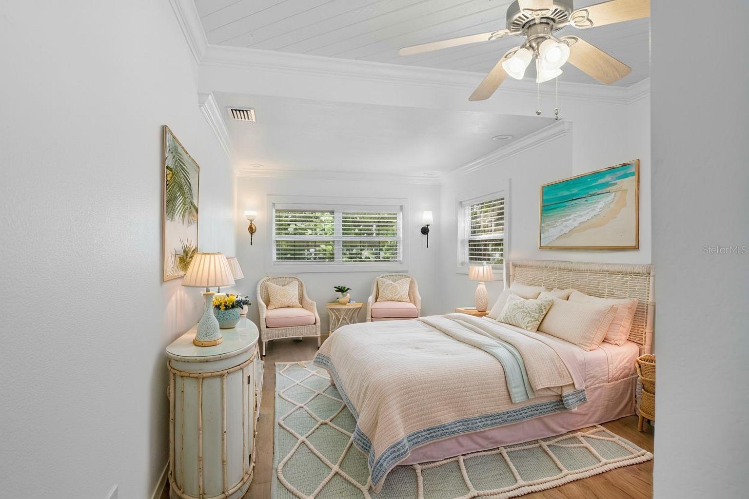 Owner's bedroom is a bright and airy retreat with multiple windows, crown molding, and wood-look flooring, offering a serene view of the tropical landscaping.