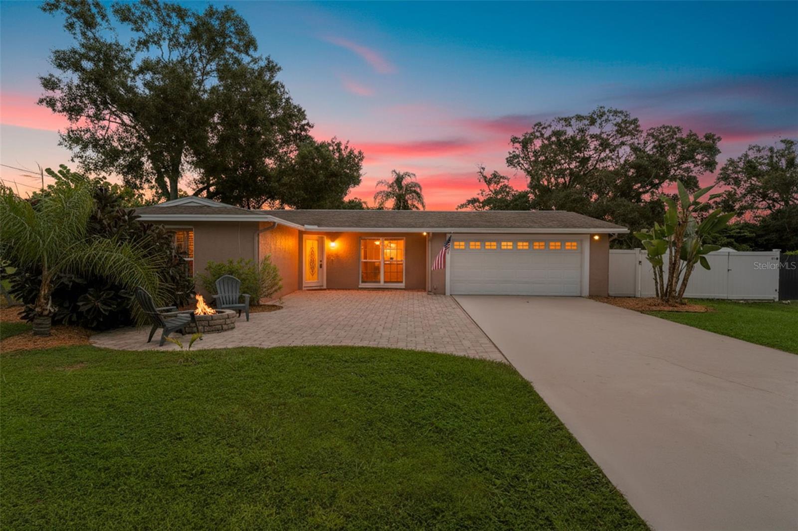 A warm and welcoming curb appeal featuring a wide paver driveway, inviting front porch, and cozy fire pit seating — all beautifully framed by mature trees and a glowing sunset backdrop.