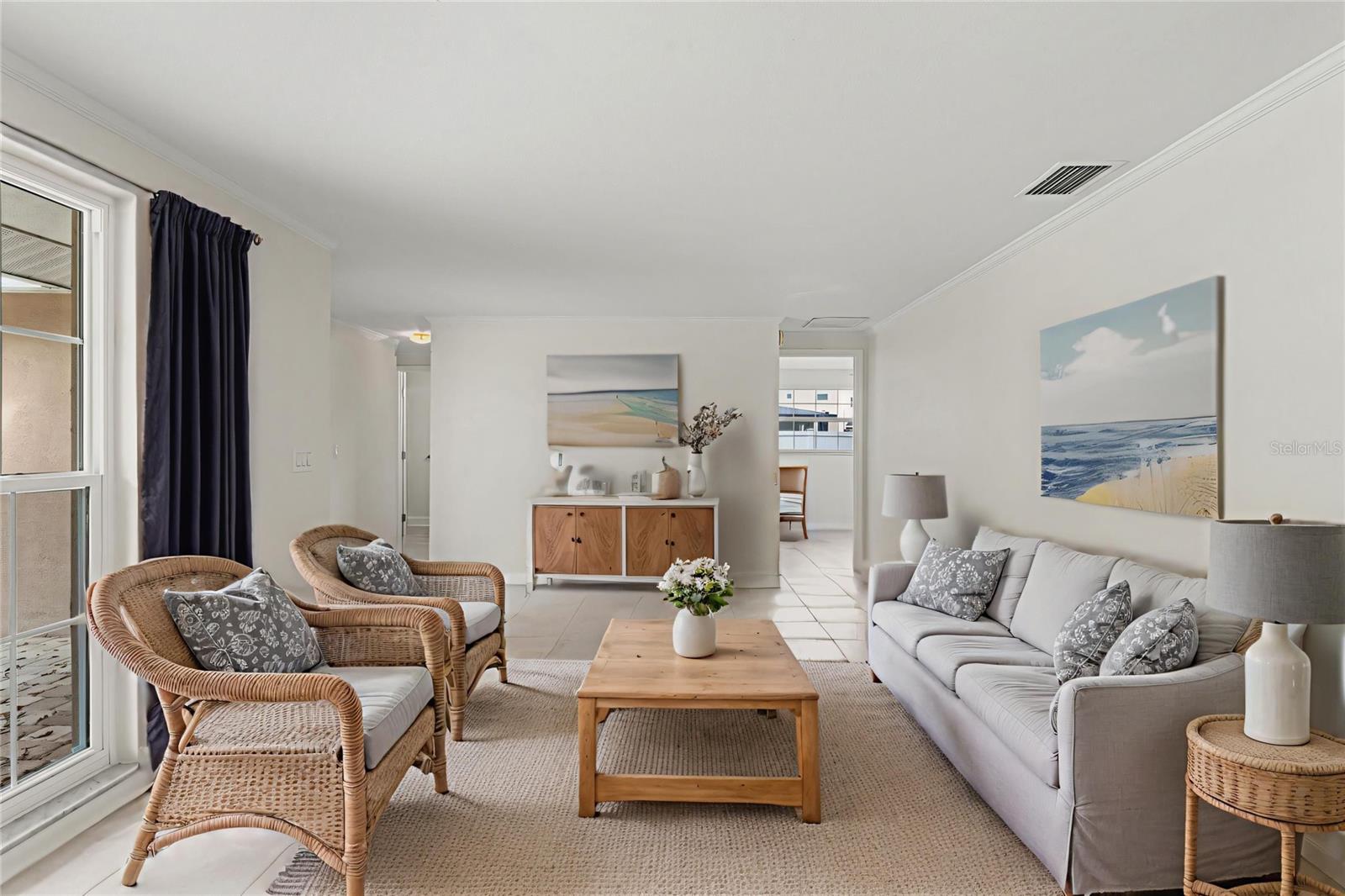 Bright and inviting living area with soft neutral tones, crown molding, and tile floors, beautifully staged with coastal-inspired décor and abundant natural light.