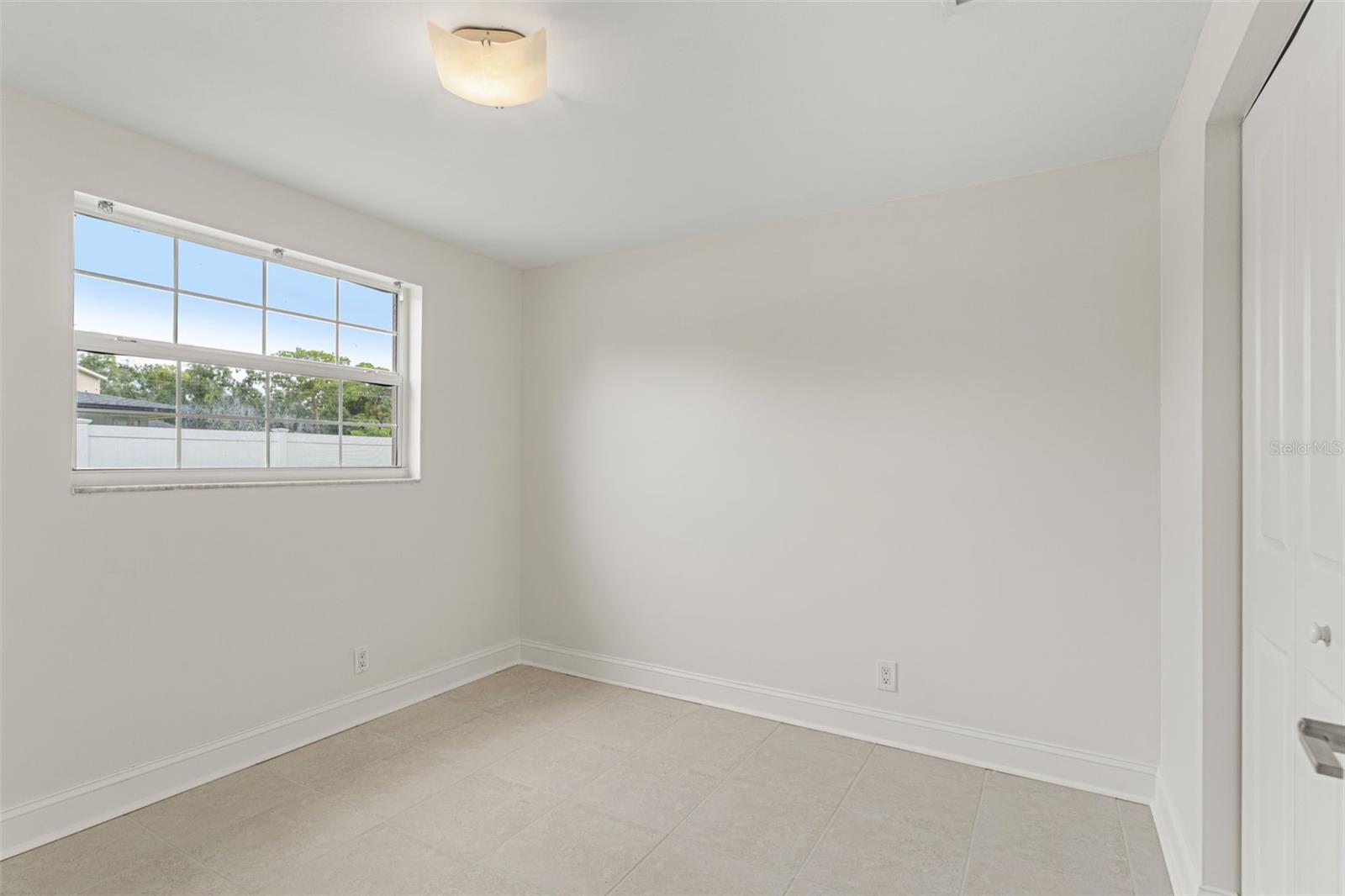 Bright guest bedroom with neutral tile floors, soft wall tones, and a large window offering plenty of natural light.