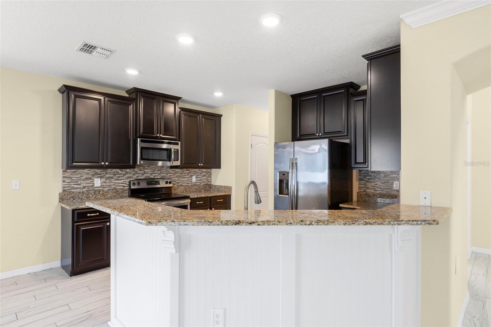 Upgraded kitchen with granite counters, tile backsplash, multi-level solid wood cabinets, stainless steel appliances, breakfast bar with wainscoting details, pantry